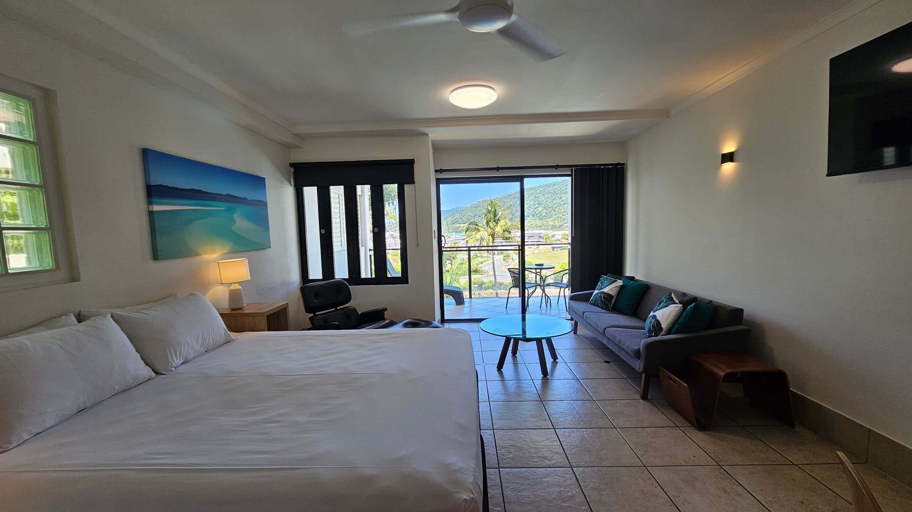 A hotel room with a large bed on the left, topped with white linens and pillows. A bedside table with a lamp is next to the bed. Above, a landscape painting hangs on the wall. In the centre, a glass coffee table is in front of a grey sofa adorned with cushions. On the right, a wall-mounted TV is visible. The room has tiled flooring and a ceiling fan. Large sliding glass doors lead to a balcony with outdoor furniture, offering a view of palm trees and distant hills.