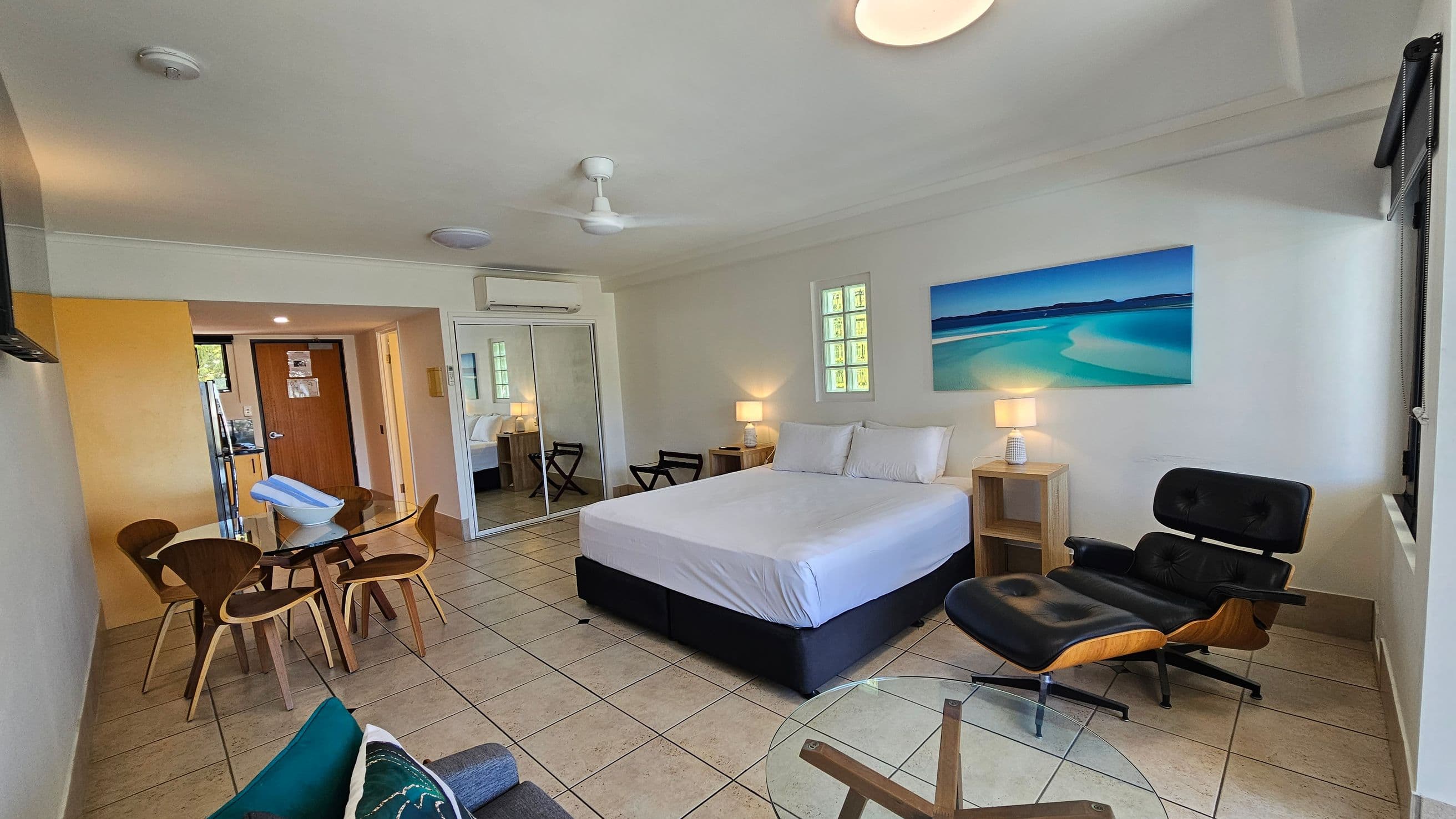 A spacious room with a large bed in the centre, flanked by two bedside tables with lamps. On the right, there's a black lounge chair and a window. A vibrant beach painting hangs above the bed. On the left, a round glass dining table with wooden chairs is near a wooden door. The floor is tiled, and the ceiling has a fan and lights. A mirrored wardrobe is visible near the bed.