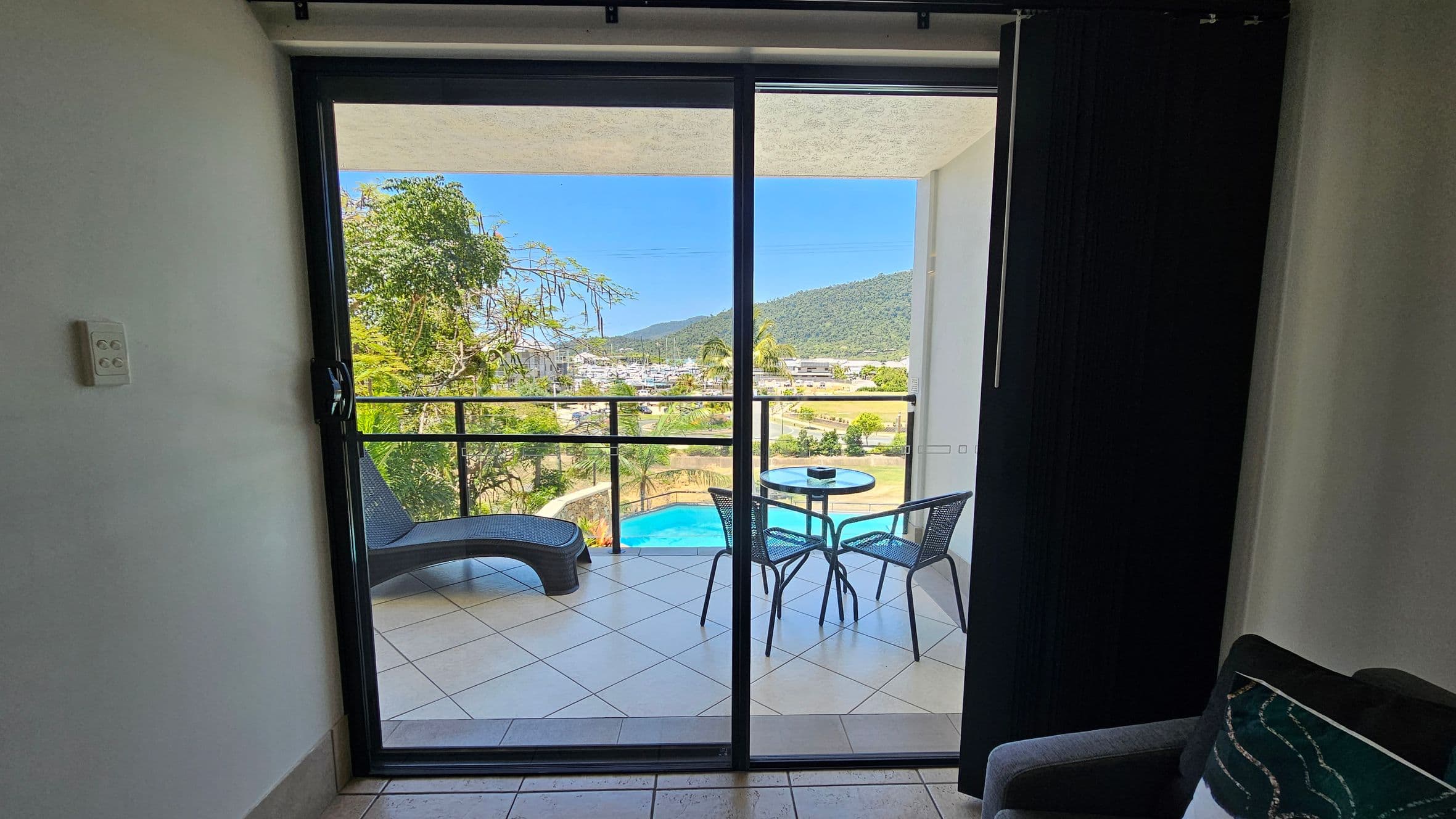 A room with a sliding glass door leading to a balcony. On the balcony, there's a round table with two chairs and a lounge chair to the left. Beyond the balcony, a view of a swimming pool, trees, and a marina with boats. In the distance, a green hill is visible. Inside, part of a grey sofa with a patterned cushion is on the right, and a light switch is on the left wall. The floor is tiled.