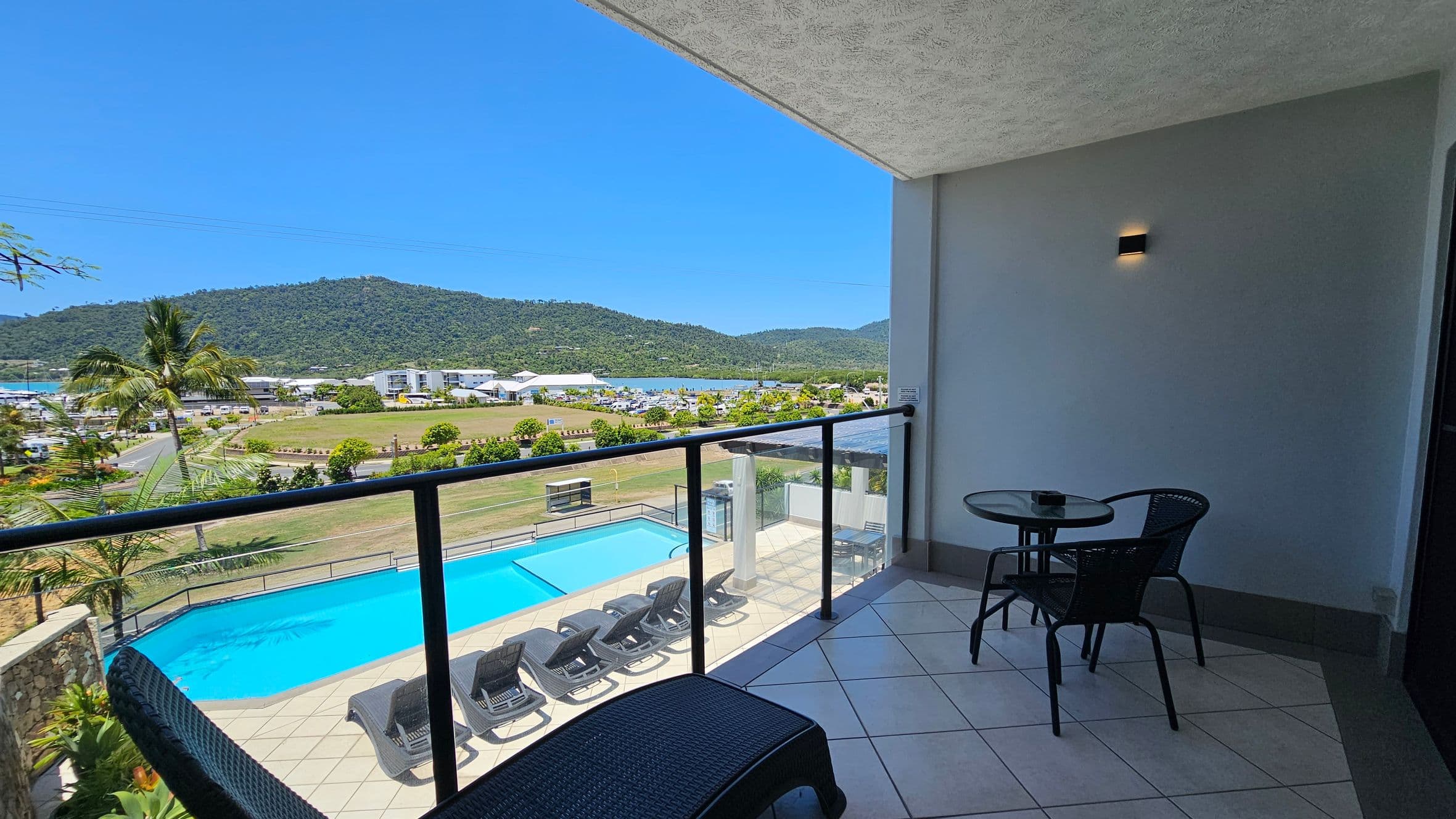 A balcony with a glass railing overlooks a swimming pool with several sun loungers. Beyond the pool, there's a grassy area with a few trees and a road. In the distance, there are buildings and a marina, with green hills in the background. On the balcony, there's a small round table with two chairs. The floor is tiled, and the ceiling is textured.
