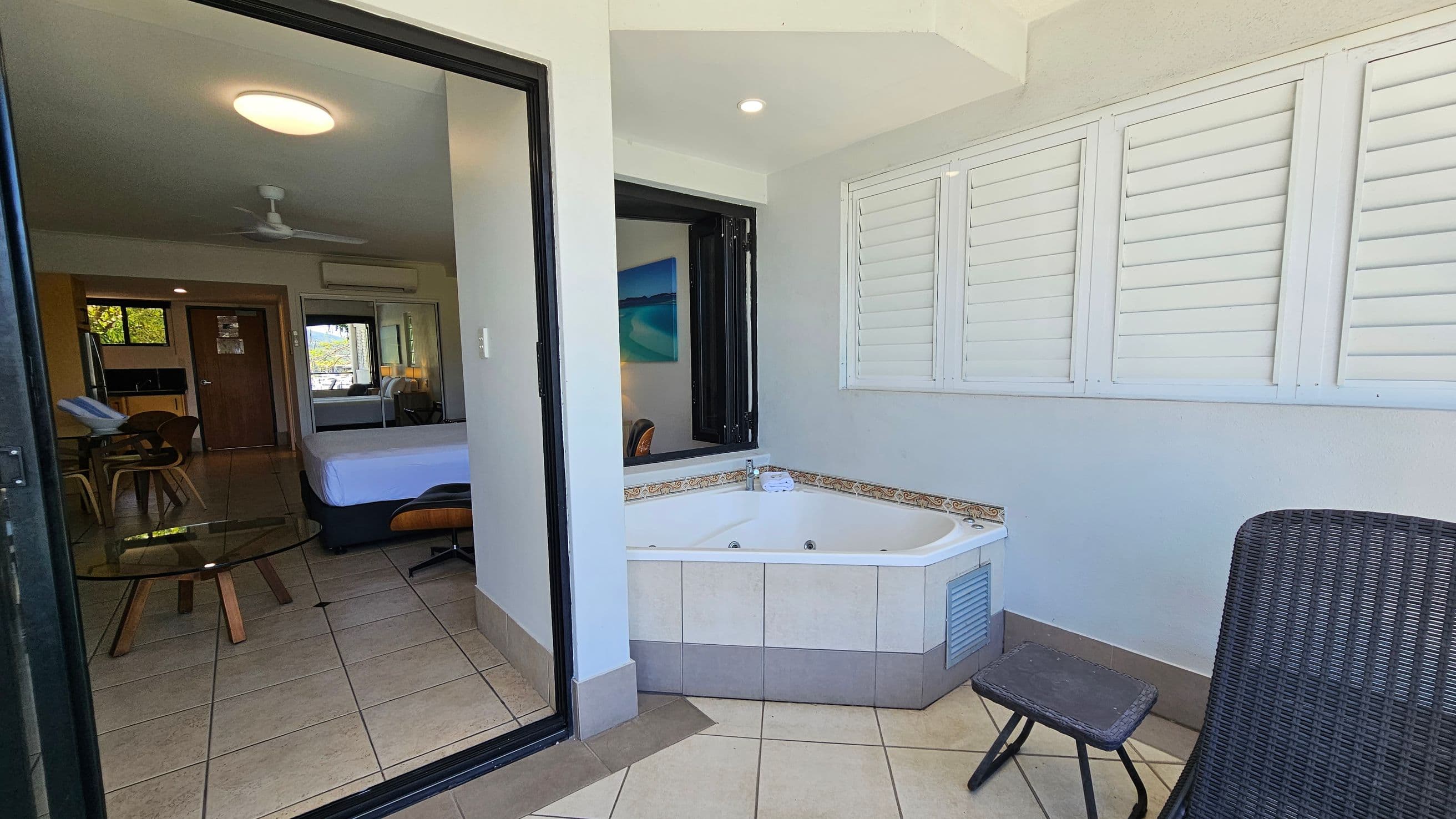A room with a tiled floor features a corner jacuzzi tub with decorative trim on the right, next to white louvred shutters. A small wicker chair and footstool are nearby. On the left, a sliding glass door opens to a room with a bed, a glass-topped table, and wooden chairs. A ceiling fan and light fixture are visible, along with a kitchenette area and a closed wooden door. A painting hangs on the wall above the bed.
