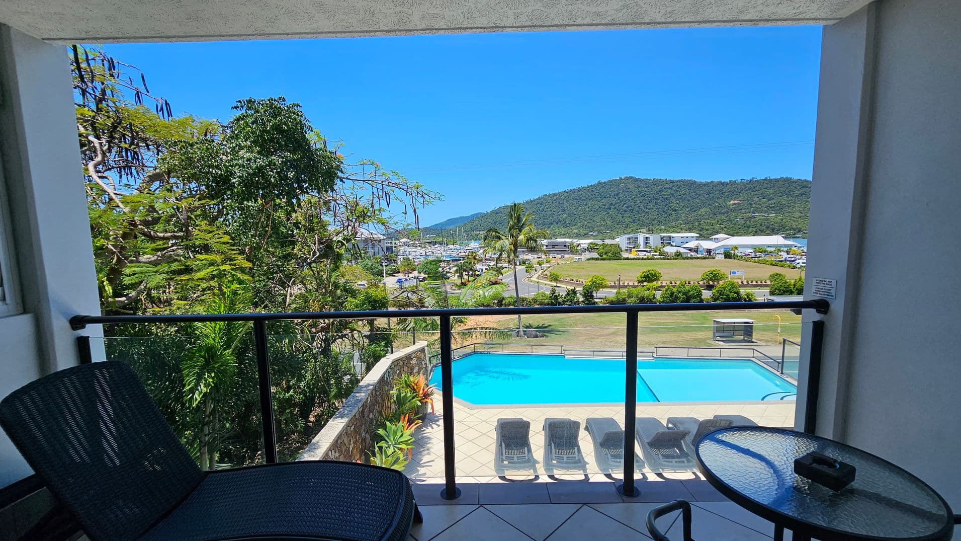 A view from a balcony with a black railing. On the left, there is a black lounge chair. In the foreground, a round glass table with an ashtray is visible. Below, a swimming pool is surrounded by a tiled area with several sun loungers. In the background, there are trees, a road, and a marina with boats. A hill with greenery is visible in the distance.