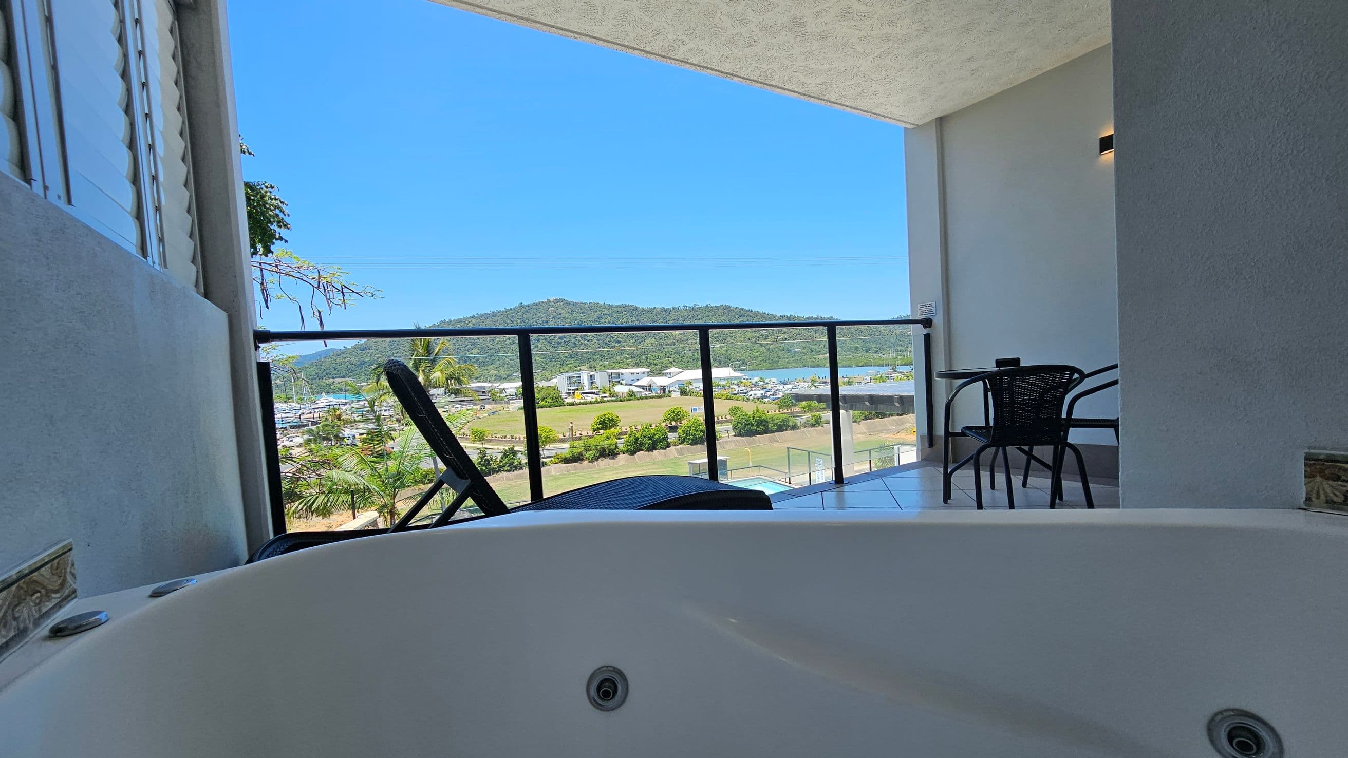 A balcony view with a white jacuzzi in the foreground. To the right, there is a small table with two black chairs. The balcony has a glass railing, offering a view of green hills and a marina with boats in the distance. The floor is tiled, and the ceiling is light-coloured. On the left, there are louvred windows partially visible.