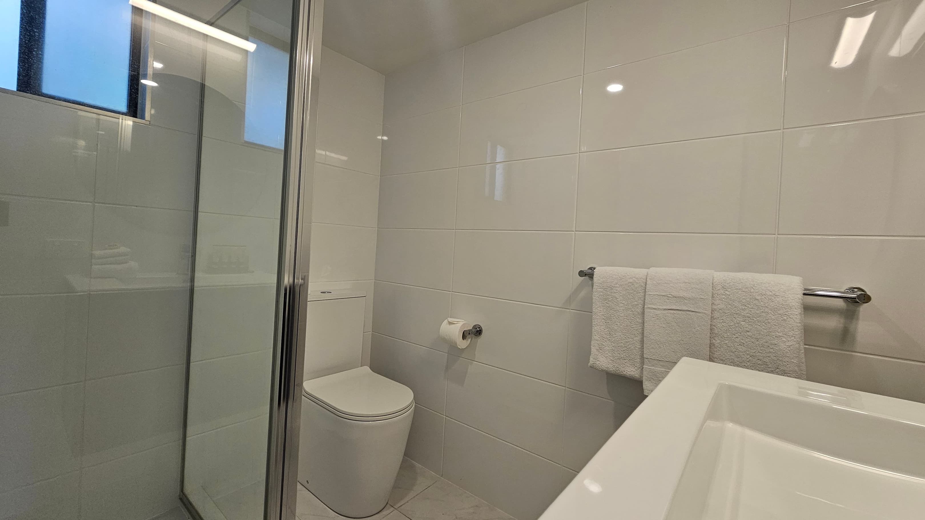 A bathroom with light-coloured tiled walls. On the left, a white sink is partially visible above a chrome towel rail holding a folded towel. In the centre, a toilet paper holder with a roll is mounted on the wall. On the right, a glass shower enclosure with a metal frame reflects light. The ceiling has recessed lighting, and the floor is tiled.
