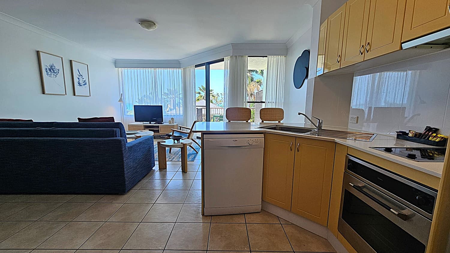 A modern living space with a kitchen on the right featuring wooden cabinets, a sink, and an oven. A dishwasher is below the counter. On the left, a blue sofa faces a TV on a wooden stand. Two framed artworks hang on the wall above the sofa. In the centre, a small round table and two bar stools are near a large window with sheer curtains, offering a view of palm trees. The floor is tiled, and the ceiling has a simple light fixture.