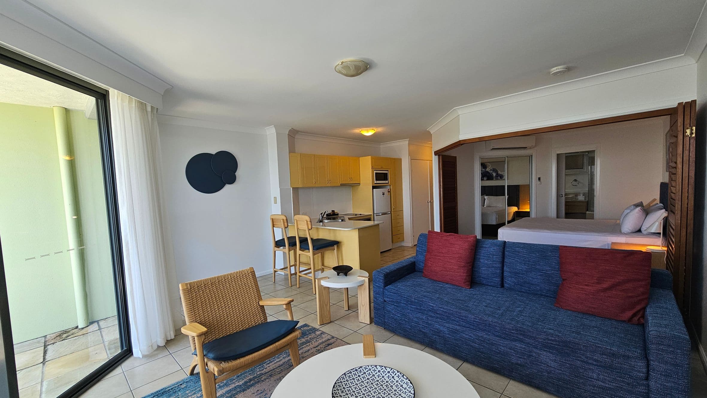 A modern living space with a sliding glass door on the left, leading to a balcony. Inside, there's a wicker chair and a blue sofa with red cushions. A round coffee table with a decorative bowl sits in front. The kitchen area is in the background, featuring light wood cabinets, a small island with two bar stools, and appliances. On the right, a bedroom area with a bed and mirrored wardrobe is partially visible. The floor is tiled, and the ceiling has recessed lighting.