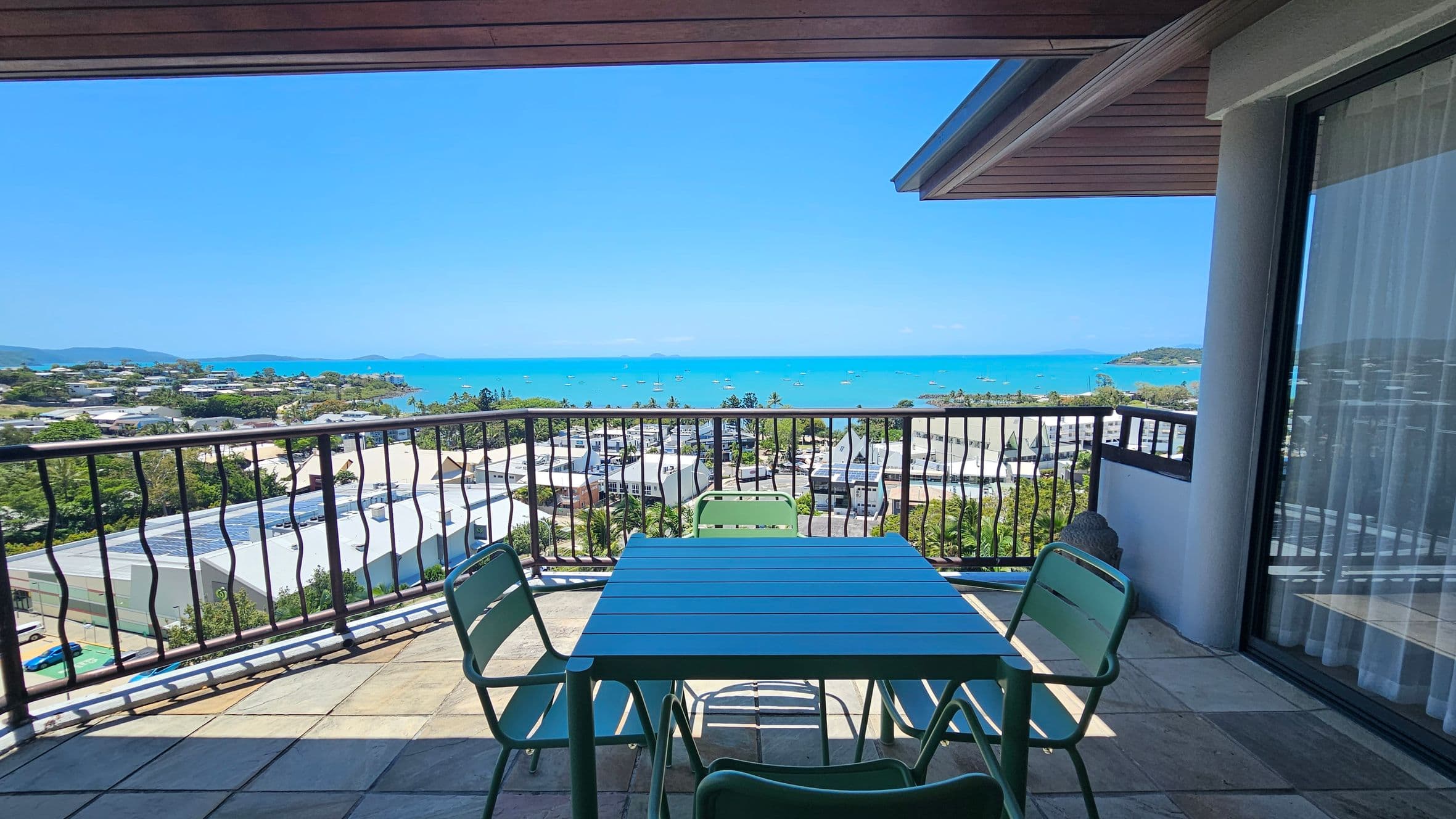 A balcony with a tiled floor features a green metal table and chairs. The railing has a wavy design, overlooking a coastal town with buildings and greenery. In the distance, a bright blue sea is dotted with small boats. The right side shows a glass door with curtains, and the ceiling has a wooden finish.