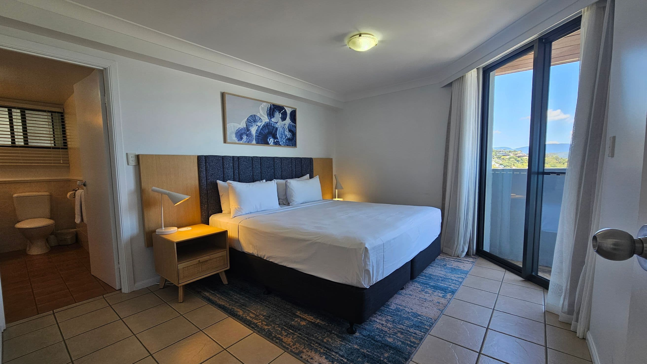A bedroom with a large bed in the centre, featuring white bedding and a dark headboard. On the left, there's a wooden bedside table with a white lamp. Above the bed hangs a framed artwork with blue circular patterns. To the left, an open door leads to a bathroom with a toilet and window blinds. On the right, glass sliding doors with sheer curtains open to a balcony with a view of greenery and hills. The floor is tiled, and the ceiling has a central light fixture.