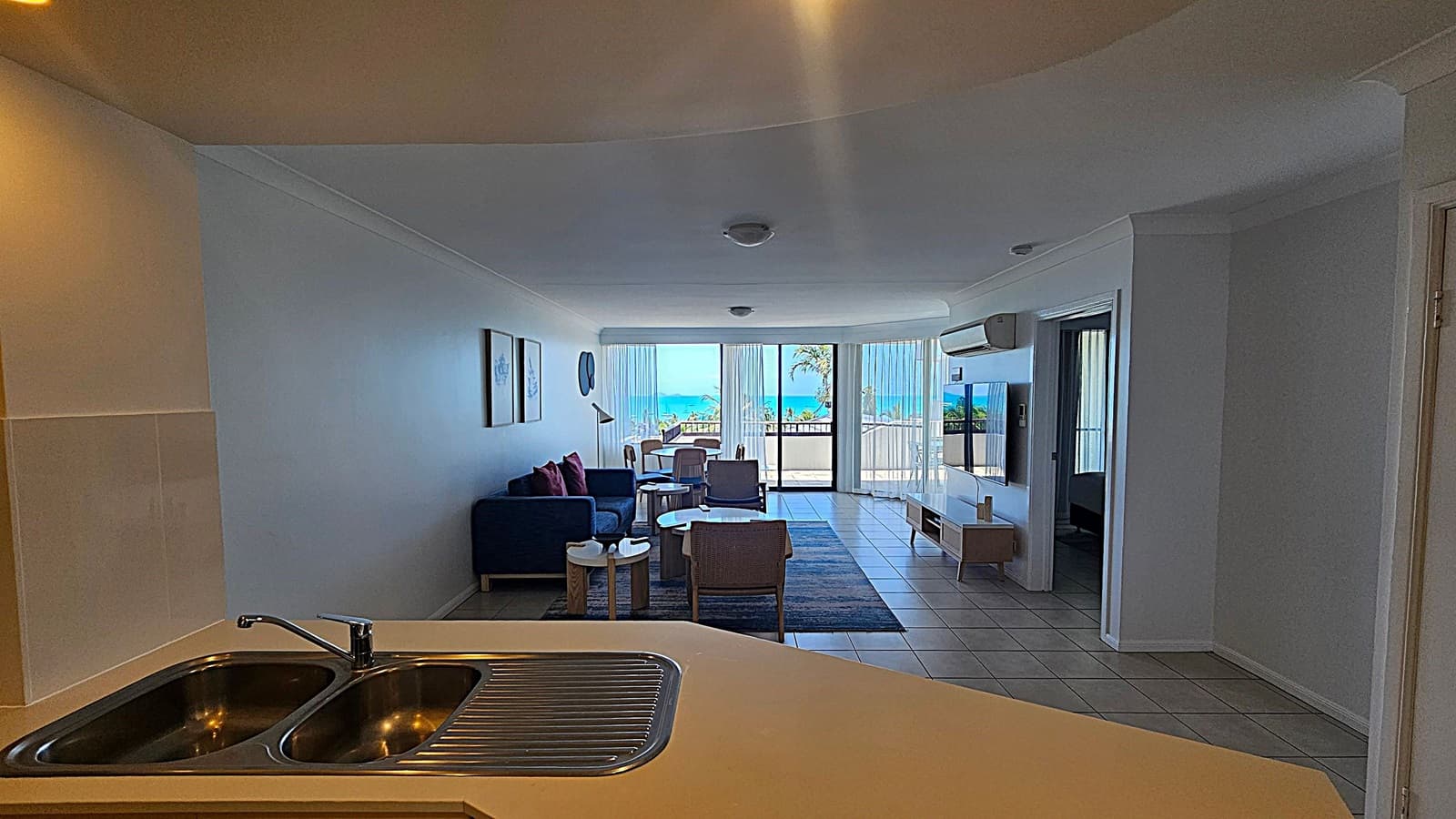 Open-plan living space with a kitchen on the left featuring light wood cabinets, an oven, and a sink. The floor is tiled. In the middle, there's a dining area with a table and chairs, and a lounge area with a dark sofa and a couple of chairs. The wall has framed artwork. On the right, large glass doors lead to a balcony with a view of the sea. The ceiling has recessed lighting, and an air conditioning unit is mounted on the wall.