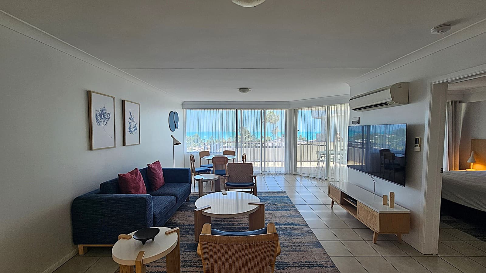 A modern living room with a tiled floor and a white ceiling. On the left, there's a dark blue sofa with red cushions, accompanied by two framed artworks on the wall. In the centre, a set of wooden chairs and round tables are placed on a patterned rug. The right side features a large flat-screen TV mounted on the wall above a wooden console. A doorway leads to a bedroom with a lit lamp. The background shows large windows with sheer curtains, offering a view of the sea and balcony furniture.