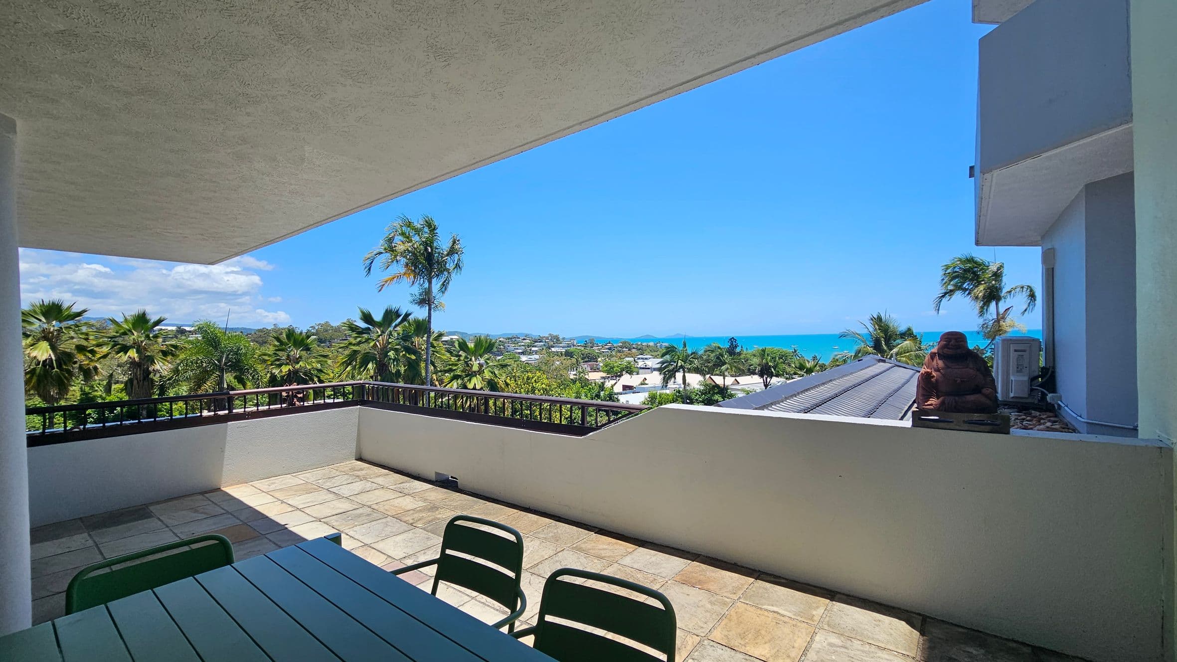 A spacious terrace with a tiled floor and a table with green chairs on the left. The view overlooks lush palm trees and a distant townscape leading to the sea. On the right, a person is sitting on a ledge next to a building with a visible air conditioning unit. The ceiling is flat and extends outward, providing shade.
