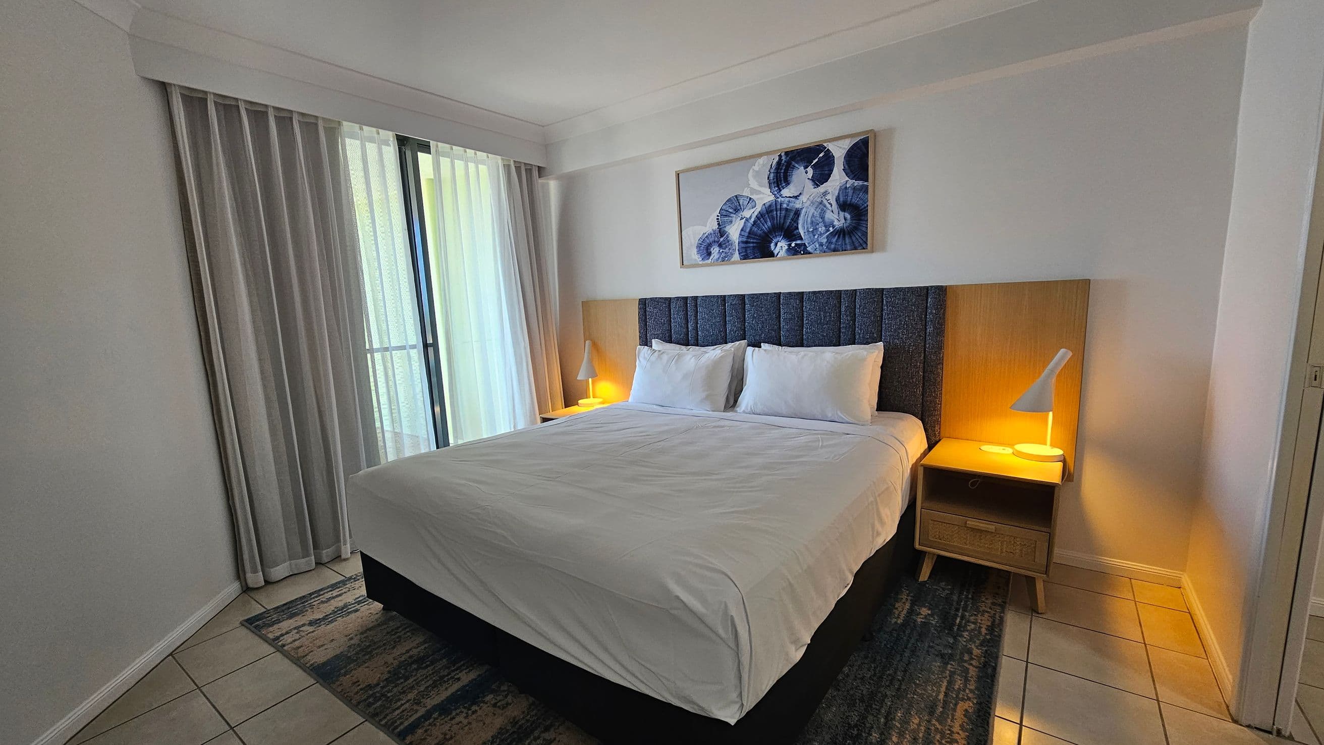 A bedroom with a large bed in the centre, covered with white linens and two pillows. On either side of the bed are wooden nightstands with modern white lamps. Above the bed hangs a framed abstract artwork. To the left, there are floor-to-ceiling windows with sheer curtains, allowing natural light to filter in. The floor is tiled, and a patterned rug is partially visible under the bed. The walls are painted white, and the ceiling is plain.