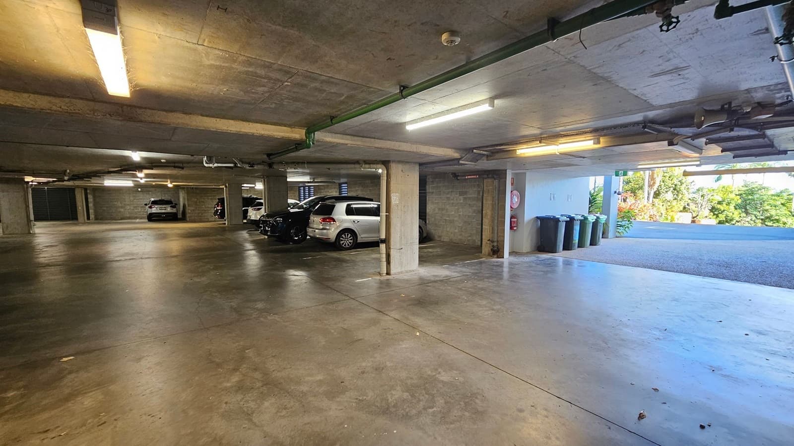 A spacious underground car park with a concrete floor and ceiling. On the left, several parked cars are visible, including a white and a black vehicle. In the middle, concrete pillars support the ceiling, with fluorescent lights overhead. On the right, an open area leads to the outside, where greenery and a road are visible. Near the entrance, there are several rubbish bins and a fire extinguisher mounted on the wall.