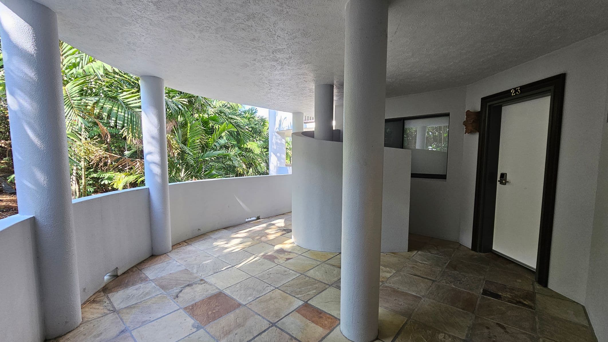 A curved corridor with a tiled floor and a white ceiling. On the left, there are large windows with views of lush green foliage outside. In the centre, white cylindrical columns support the ceiling. On the right, a door marked "23" is set in a black frame, next to a small window. The area is well-lit, with natural light streaming in from the windows.