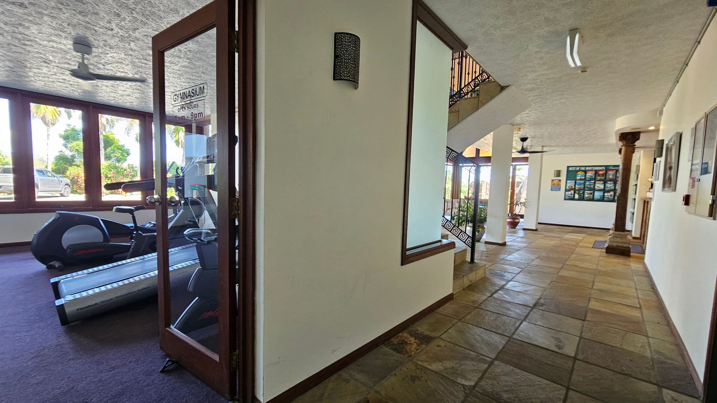 On the left, there's a gym with exercise equipment visible through a glass door. The gym has large windows with a view of trees and a parked vehicle outside. On the right, a hallway with tiled flooring leads to a staircase with decorative railings. The walls are adorned with framed pictures and a poster. A column stands near the entrance, and ceiling fans are visible above.