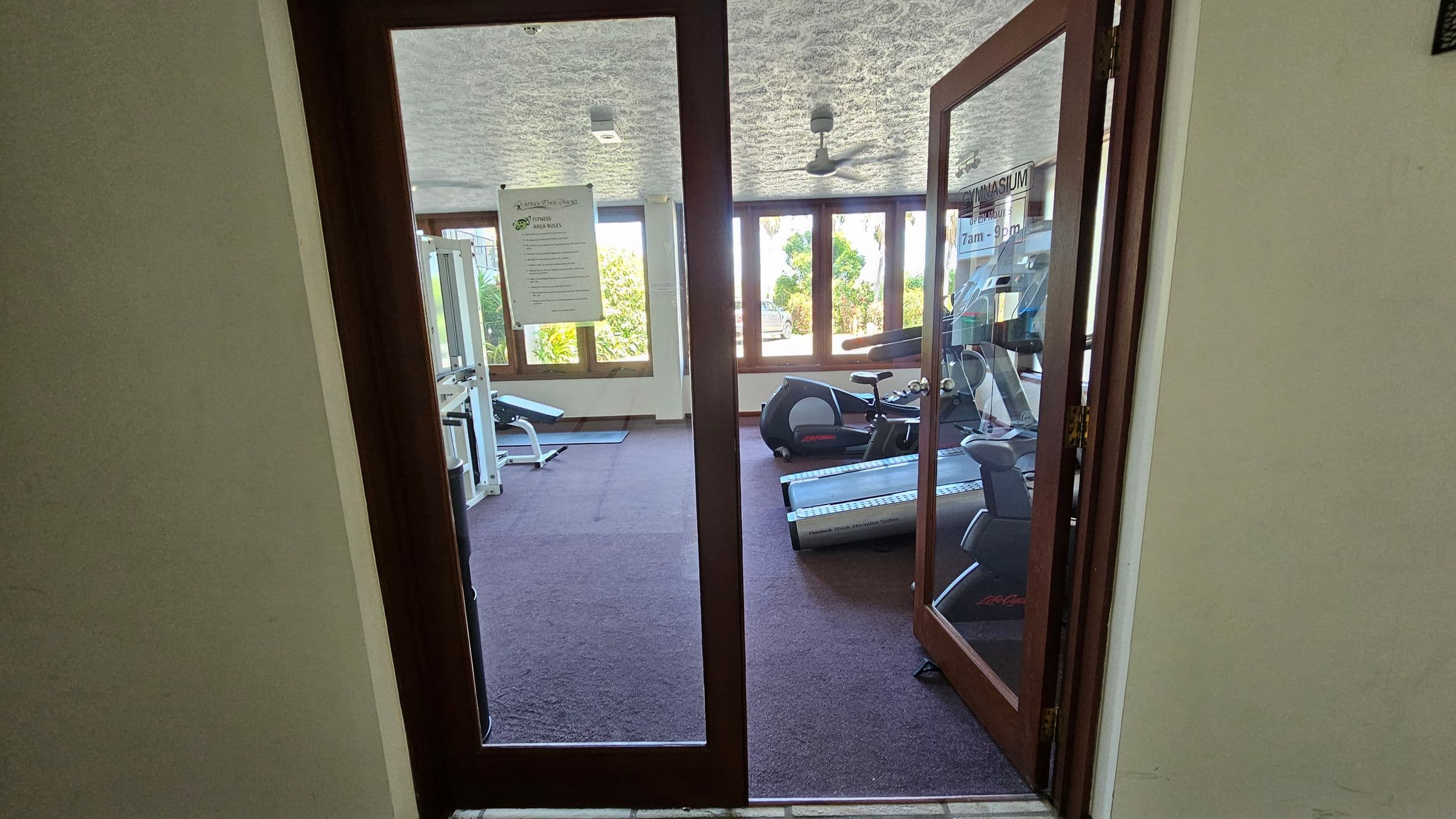 Glass doors open into a gym with a purple carpeted floor. On the left, there's a white exercise machine near large windows. On the right, a treadmill and a stationary bike are visible. The ceiling is textured with a fan in the centre. A sign with gym rules is posted on the left door, and another sign with opening hours is on the right door. Bright natural light comes through the windows, revealing greenery outside.