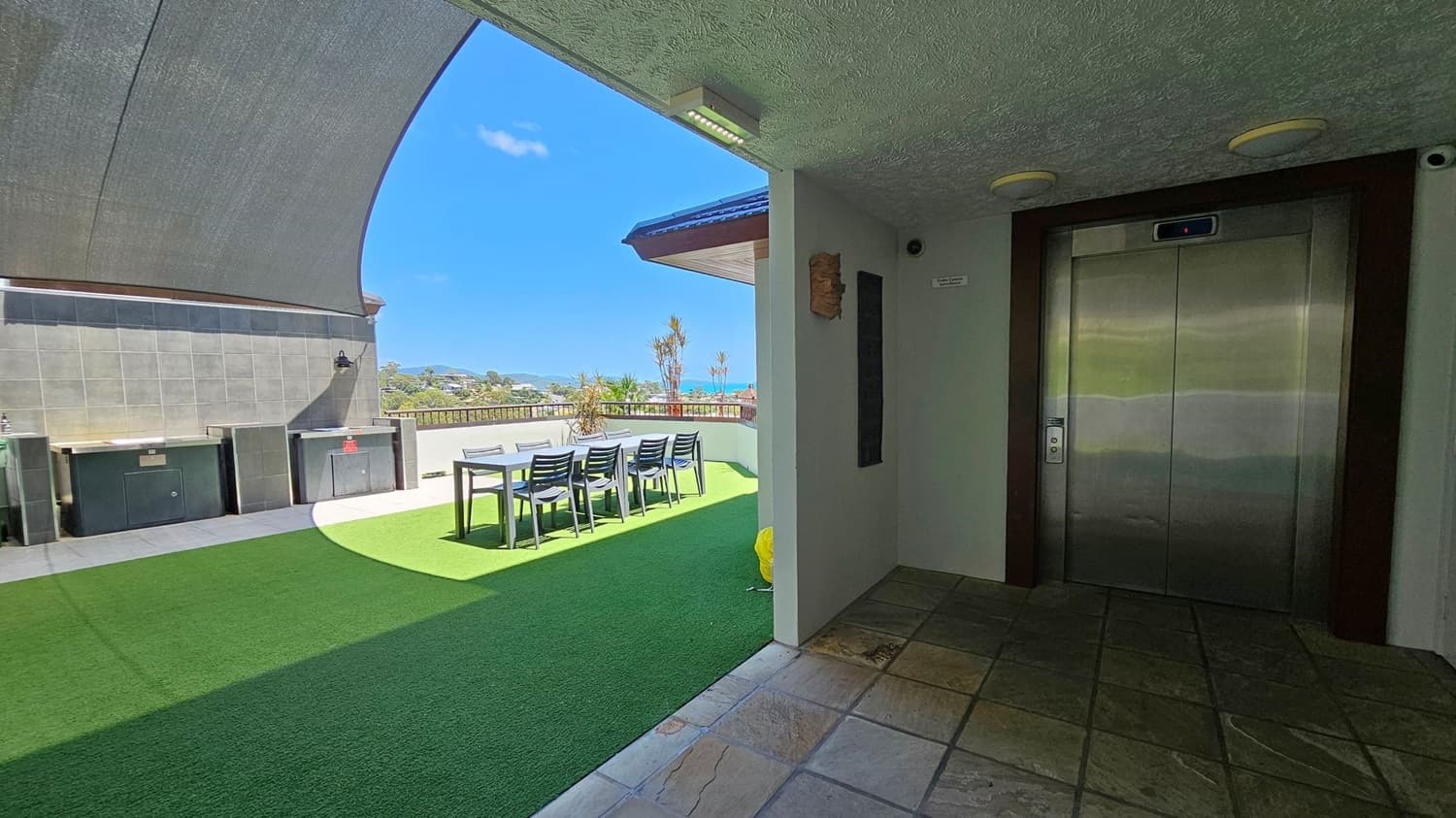 On the right, there is a metallic lift door set in a tiled wall. The floor in front of the lift is made of square tiles. On the left, a shaded outdoor area features a green artificial grass floor, a long table with several chairs, and a barbecue setup against a tiled wall. In the background, there is a view of distant hills and the sea, with a few small trees and a railing along the edge of the terrace.