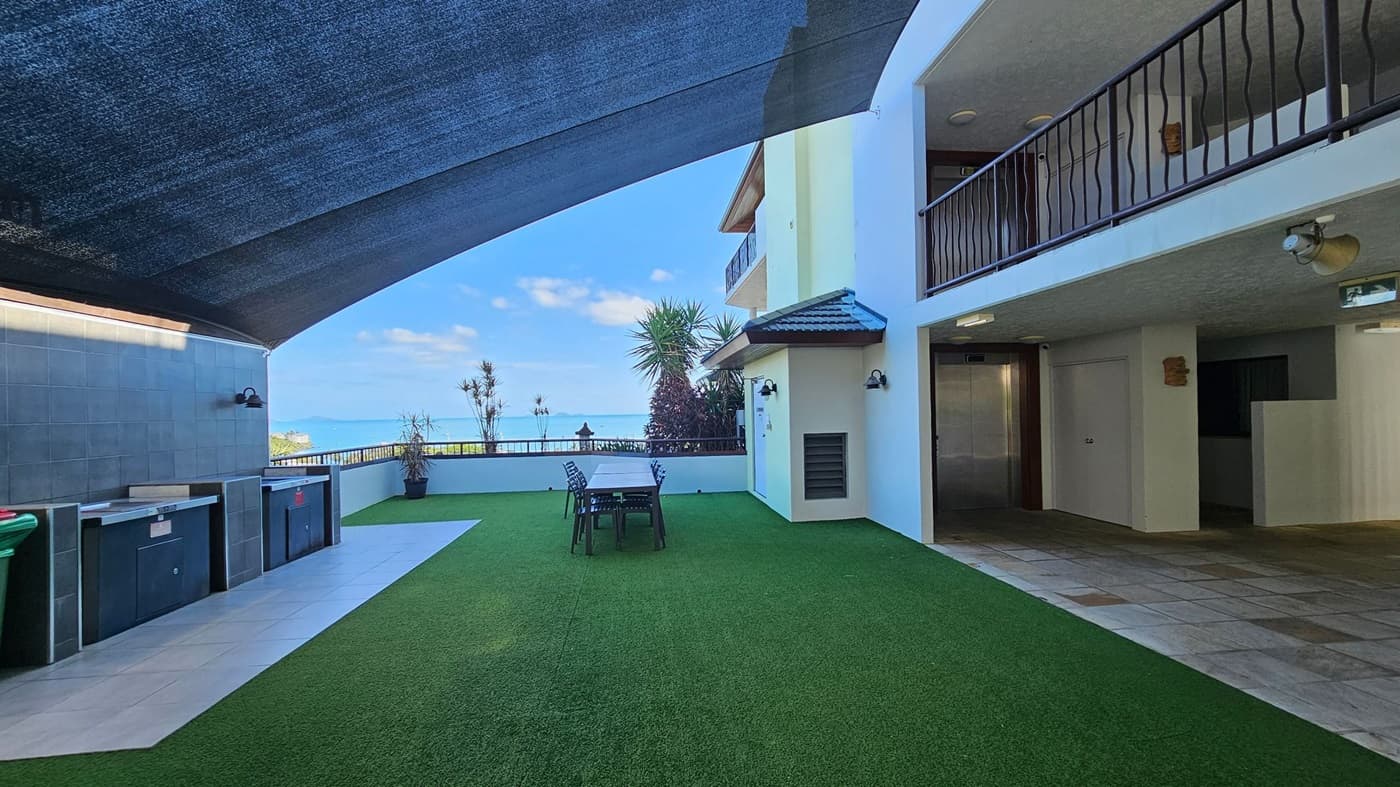 A spacious outdoor area with artificial grass covering the ground. On the left, there are built-in outdoor kitchen units with a tiled backsplash and a green bin. In the centre, a long table with chairs is set up for dining. The right side features a building with a balcony and a tiled floor area leading to an entrance. Above, a large shade cloth provides cover. In the background, potted plants and a view of the sea are visible.