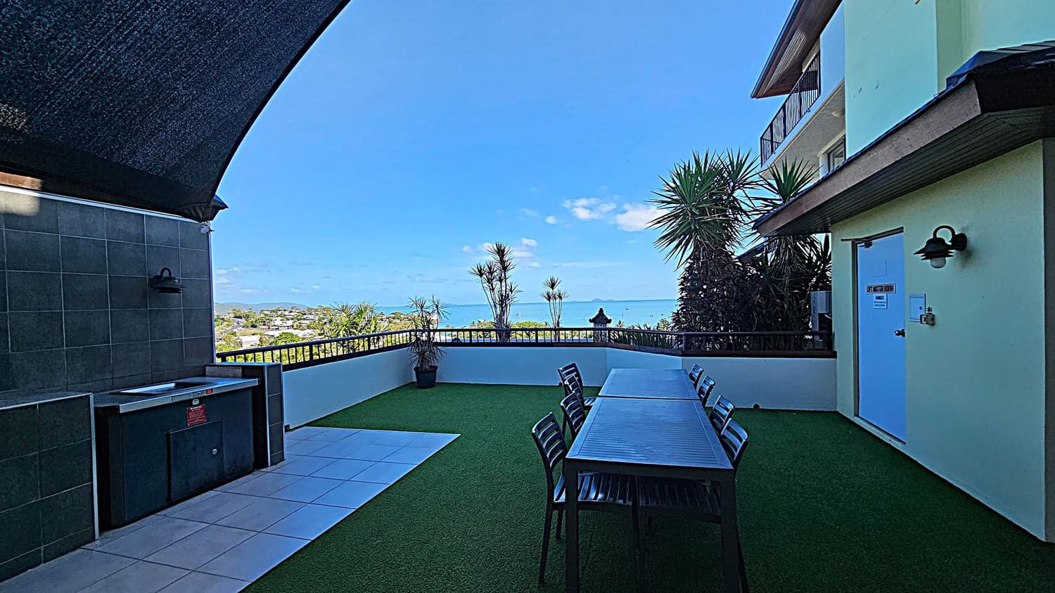 A rooftop terrace with artificial grass flooring and a long wooden table with chairs on the right. A building wall with a door labelled "Lift Motor Room" is also on the right. On the left, there's a tiled area with a barbecue grill. The background features a railing with potted plants and a view of the sea and distant landscape. A dark canopy partially covers the area.