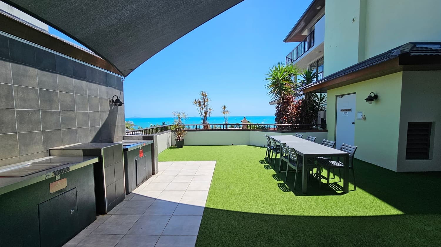 Outdoor terrace with a tiled floor on the left and artificial grass on the right. A shaded area covers stainless steel kitchen appliances against a dark tiled wall on the left. On the right, a long table with chairs is set up near a building with a white door. In the background, there are potted plants and a view of the ocean.