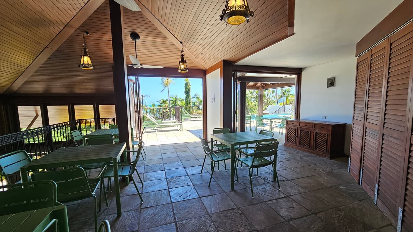 A spacious interior with a sloped wooden ceiling and hanging lanterns. On the left, large windows with wooden frames allow natural light to enter. In the centre, a black metal railing with a geometric pattern lines a staircase. On the right, there are tables and chairs with a view of greenery outside. The floor is tiled, and ceiling fans are visible.