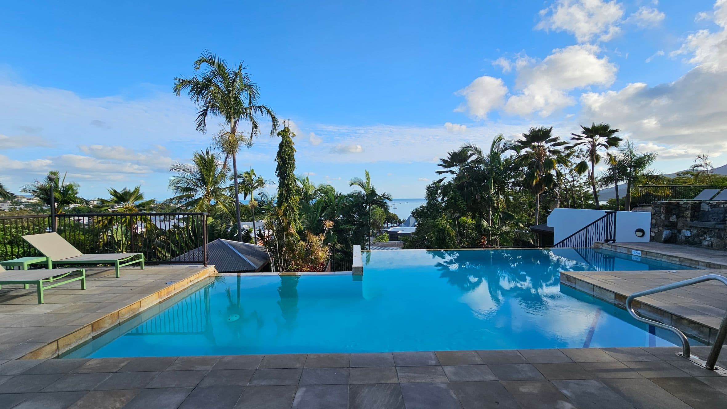 An infinity pool with clear blue water is in the centre, surrounded by a tiled deck. On the left, there are two green sun loungers. The right side features a stone wall and a metal handrail leading into the pool. In the background, lush palm trees and tropical vegetation are visible, with a glimpse of the sea in the distance.