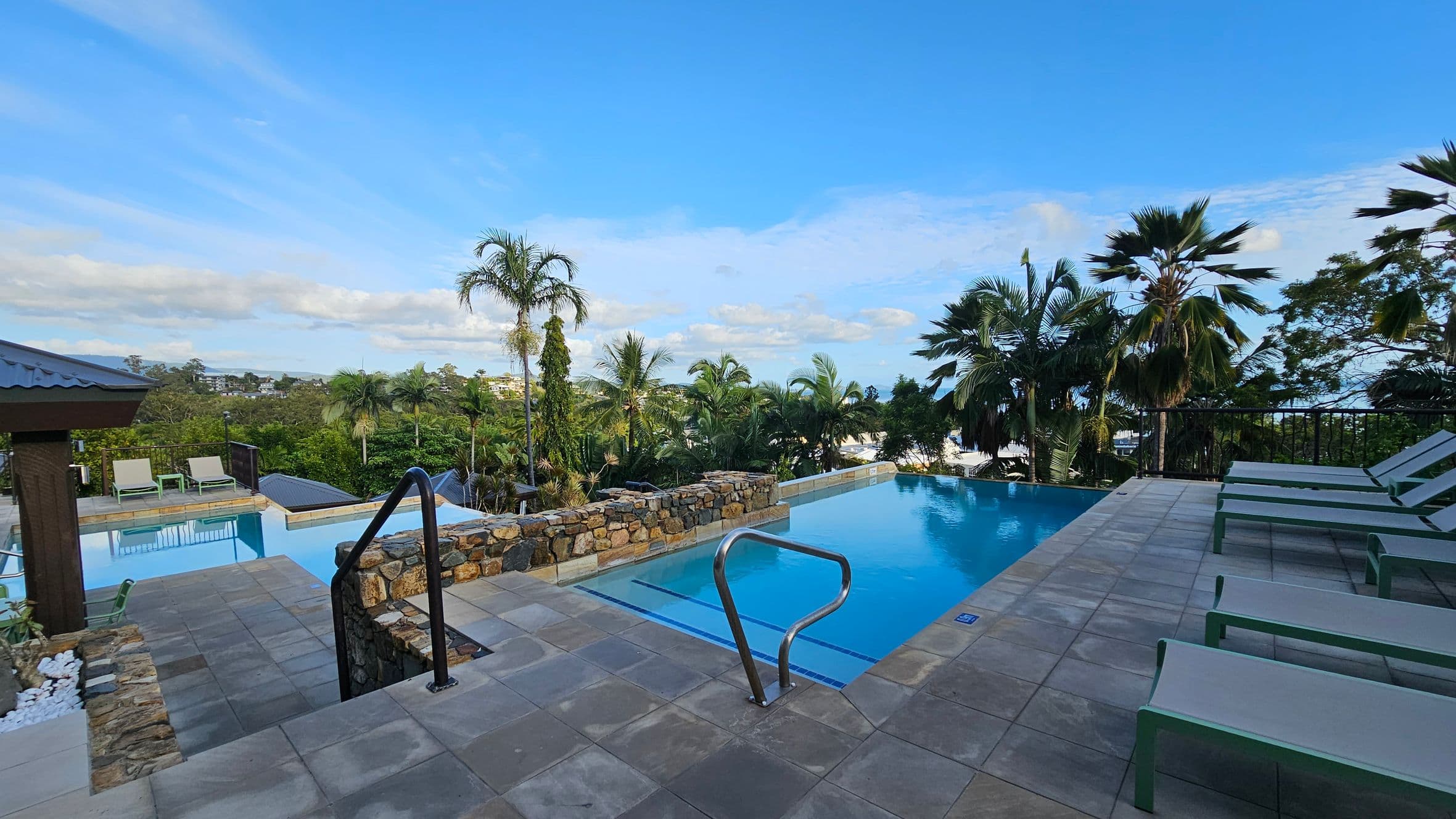 A luxurious outdoor setting with two swimming pools. The foreground features a stone-bordered pool with metal handrails. To the right, several green sun loungers are lined up on a tiled deck. In the background, lush palm trees and greenery surround the area, with a glimpse of distant buildings. The left side shows another pool with a small pavilion and more loungers.