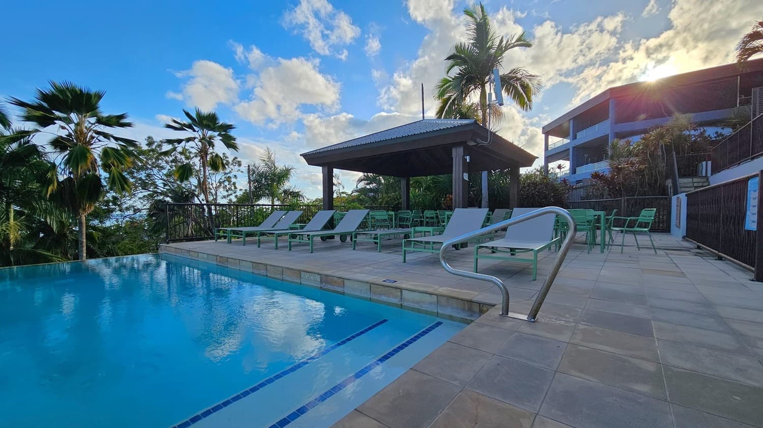 An outdoor pool with clear blue water is on the left, bordered by a tiled deck. On the right, several sun loungers and green chairs are arranged under a wooden gazebo. Palm trees are scattered around, and a modern building is visible in the background. The sun is partially hidden behind the building, casting light over the scene.