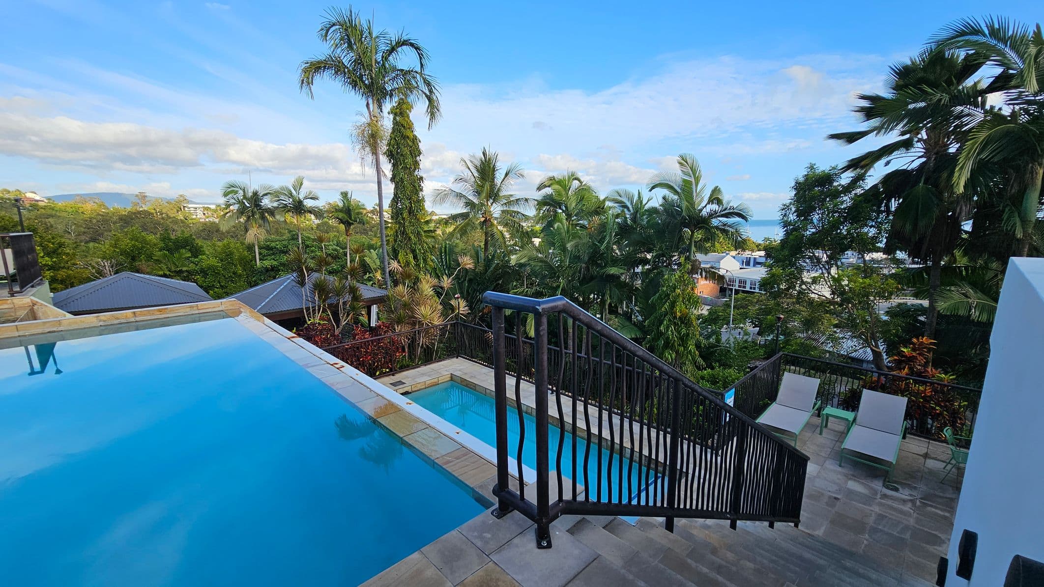 A view of a luxurious outdoor area with an infinity pool on the left, bordered by stone tiles. A staircase with a black railing descends to a lower level where there are two sun loungers and a small table. The background is filled with lush green palm trees and other tropical vegetation. Rooftops of nearby buildings are visible beyond the trees, with a glimpse of the ocean in the distance.