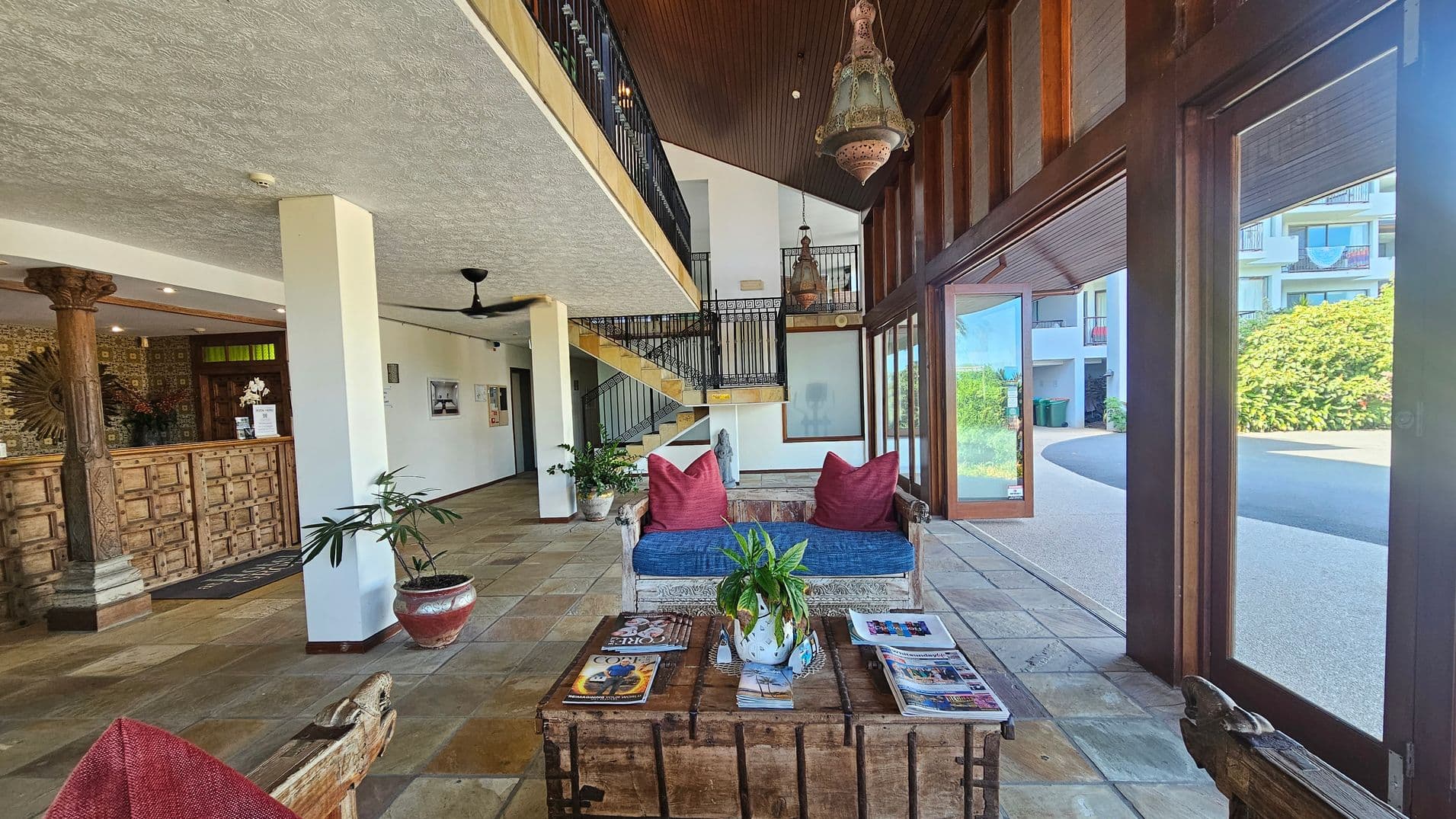 A spacious lobby with a rustic wooden reception desk on the left, adorned with intricate carvings and a decorative column. A staircase with black railings leads to an upper level. In the centre, a seating area features a wooden bench with red cushions and a blue seat, facing a wooden coffee table with magazines and a plant. Large windows on the right offer a view of greenery and a building outside. The floor is tiled, and the ceiling is textured with a fan and hanging lights.