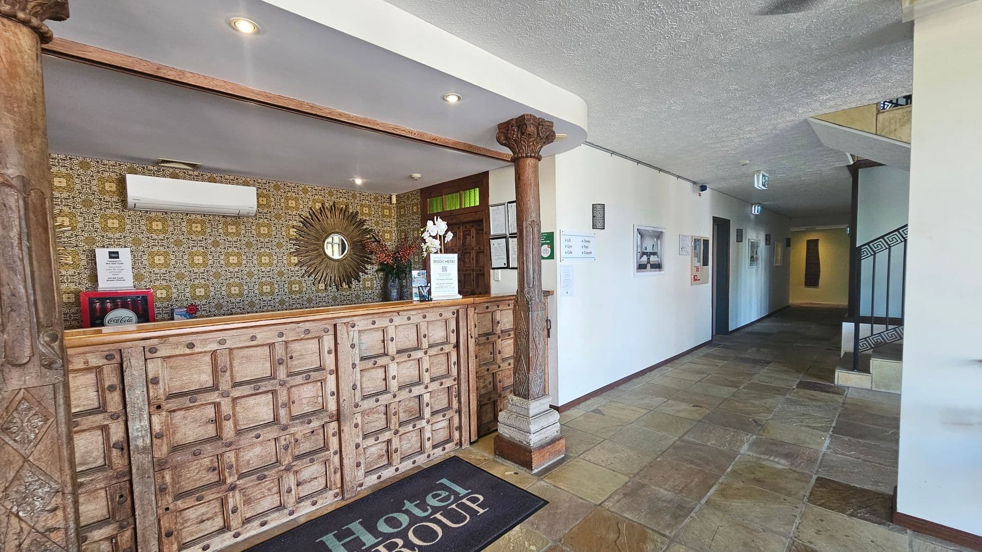 A hotel reception area with a rustic wooden counter on the left, featuring intricate carvings and a decorative pillar. Behind the counter, there's a patterned wallpaper with a sunburst mirror and an air conditioning unit. A small Coca-Cola fridge is visible. The floor is tiled with a mix of green and brown stones. On the right, a hallway leads to rooms, with framed pictures on the walls and a staircase with a decorative railing. The ceiling is white with recessed lighting.