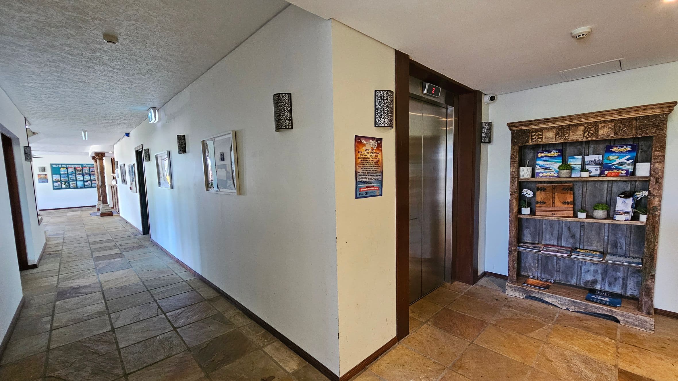 A hallway with a stone-tiled floor and a textured ceiling. On the right, there's a wooden shelf with brochures and small potted plants. Next to it is a lift with a digital display showing the number 2. The left side of the hallway has framed pictures on the wall and leads to a corridor with a column and a display board in the distance. Wall-mounted lights are evenly spaced along the walls.