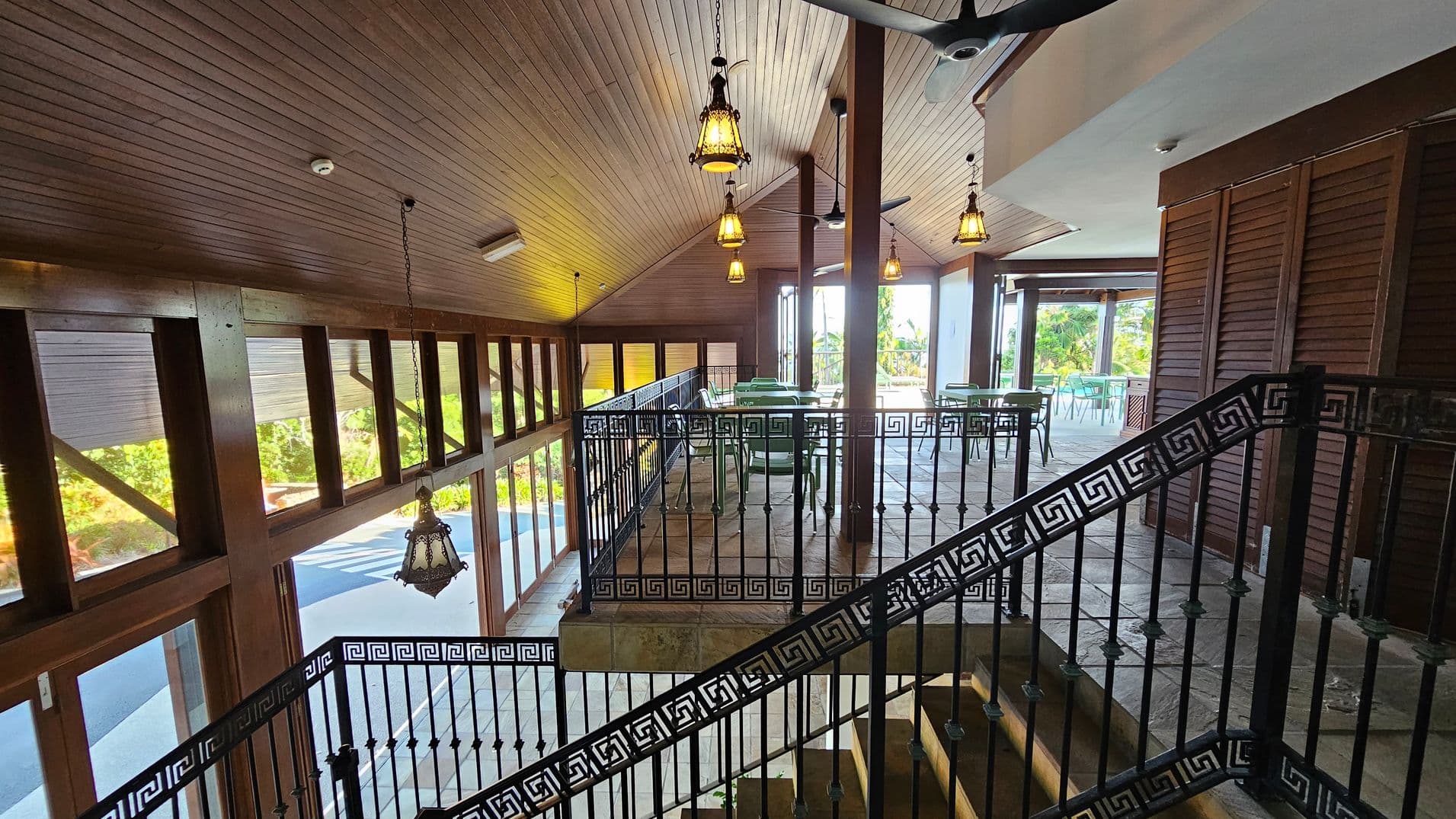 A spacious interior with a sloped wooden ceiling and hanging lanterns. On the left, large windows with wooden frames allow natural light to enter. In the centre, a black metal railing with a geometric pattern lines a staircase. On the right, there are tables and chairs with a view of greenery outside. The floor is tiled, and ceiling fans are visible.