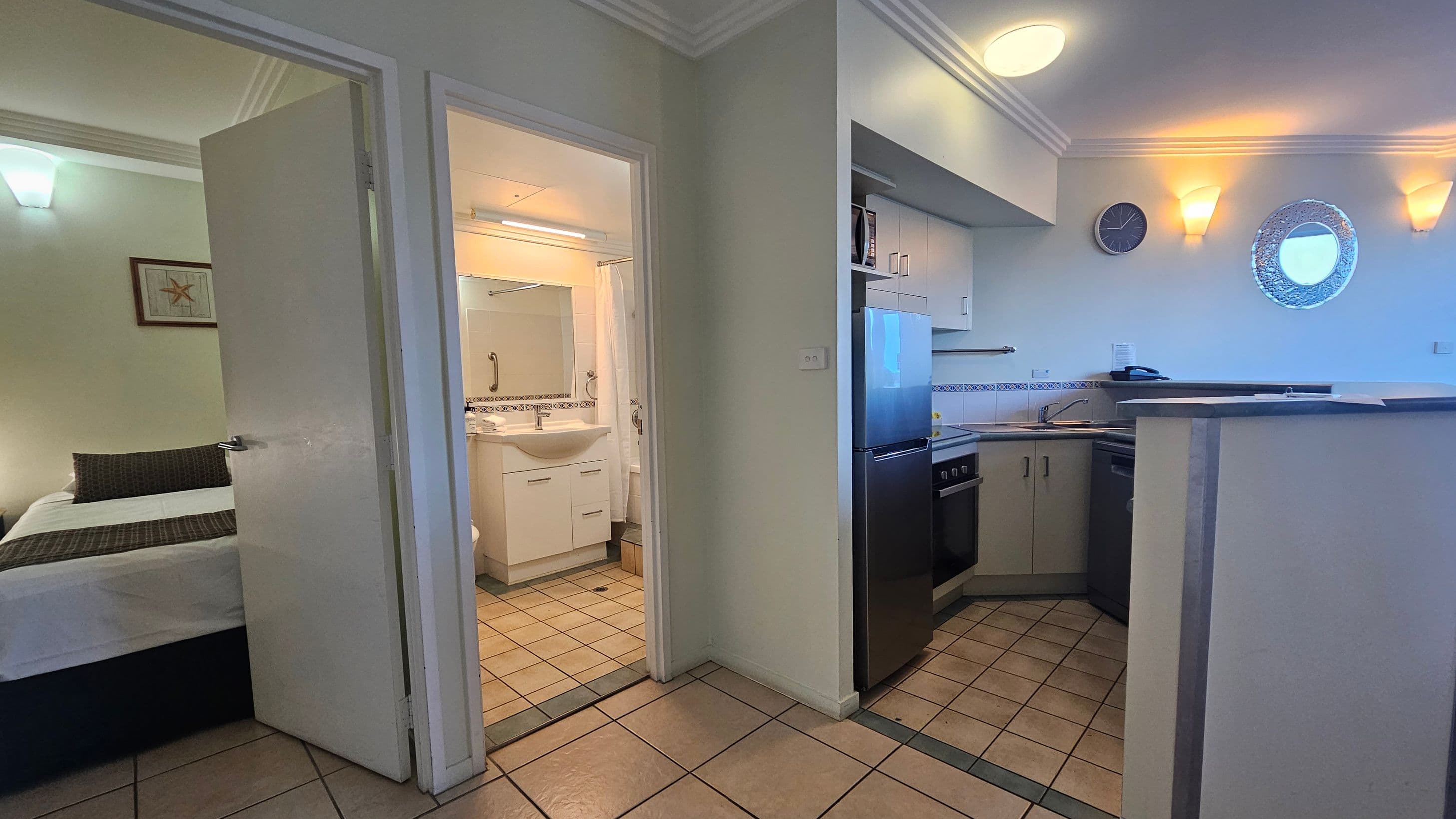 A small apartment interior with a tiled floor. On the left, there's a partially open door leading to a bedroom with a bed and a framed picture on the wall. In the centre, another door opens to a bathroom with a sink and mirror. On the right, a compact kitchen area features a fridge, oven, and sink. Above the kitchen counter, a wall clock and a decorative round mirror are mounted. The ceiling has recessed lighting and wall sconces.