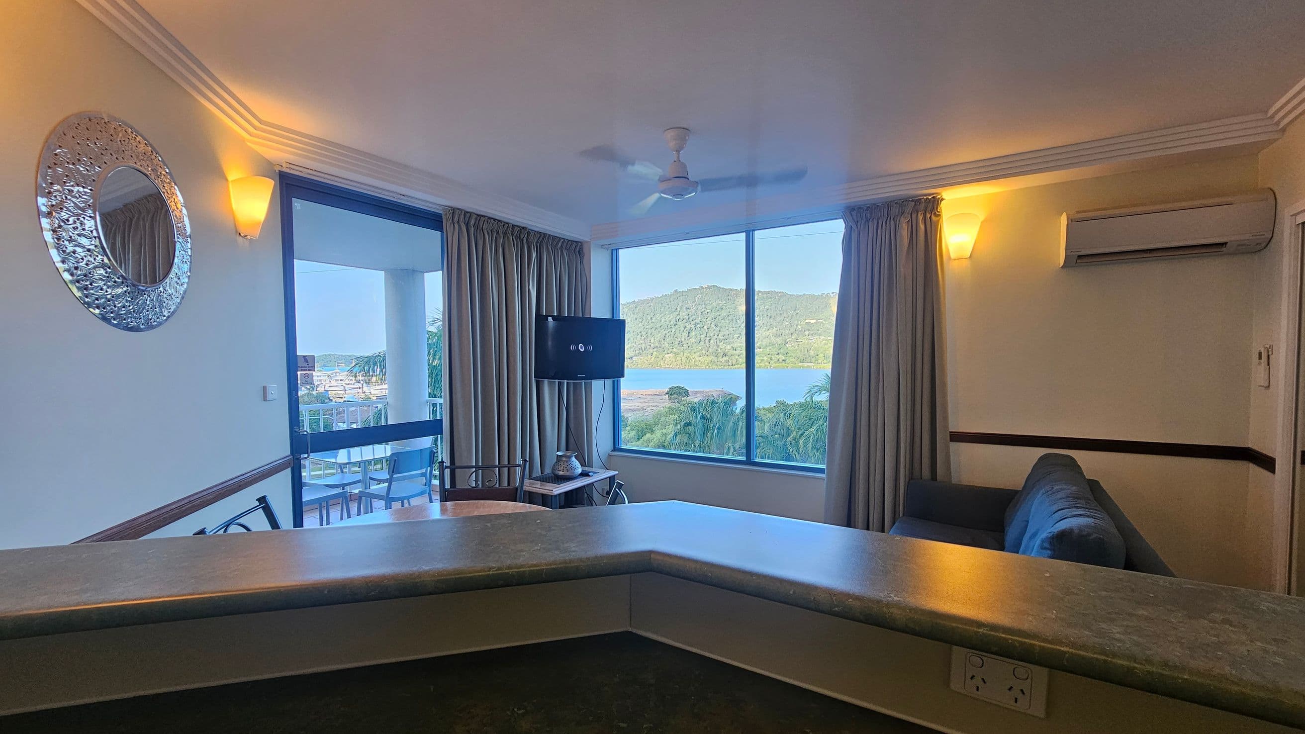 A room with a large window and sliding glass door on the left, offering a view of a balcony with outdoor furniture and a scenic landscape of hills and water. A round decorative mirror is on the left wall. In the centre, a TV is mounted near the window, and a small table with a decorative item is below it. On the right, a grey sofa sits beneath an air conditioning unit. The ceiling has a fan, and the room is lit by wall-mounted lights. A countertop is in the foreground.