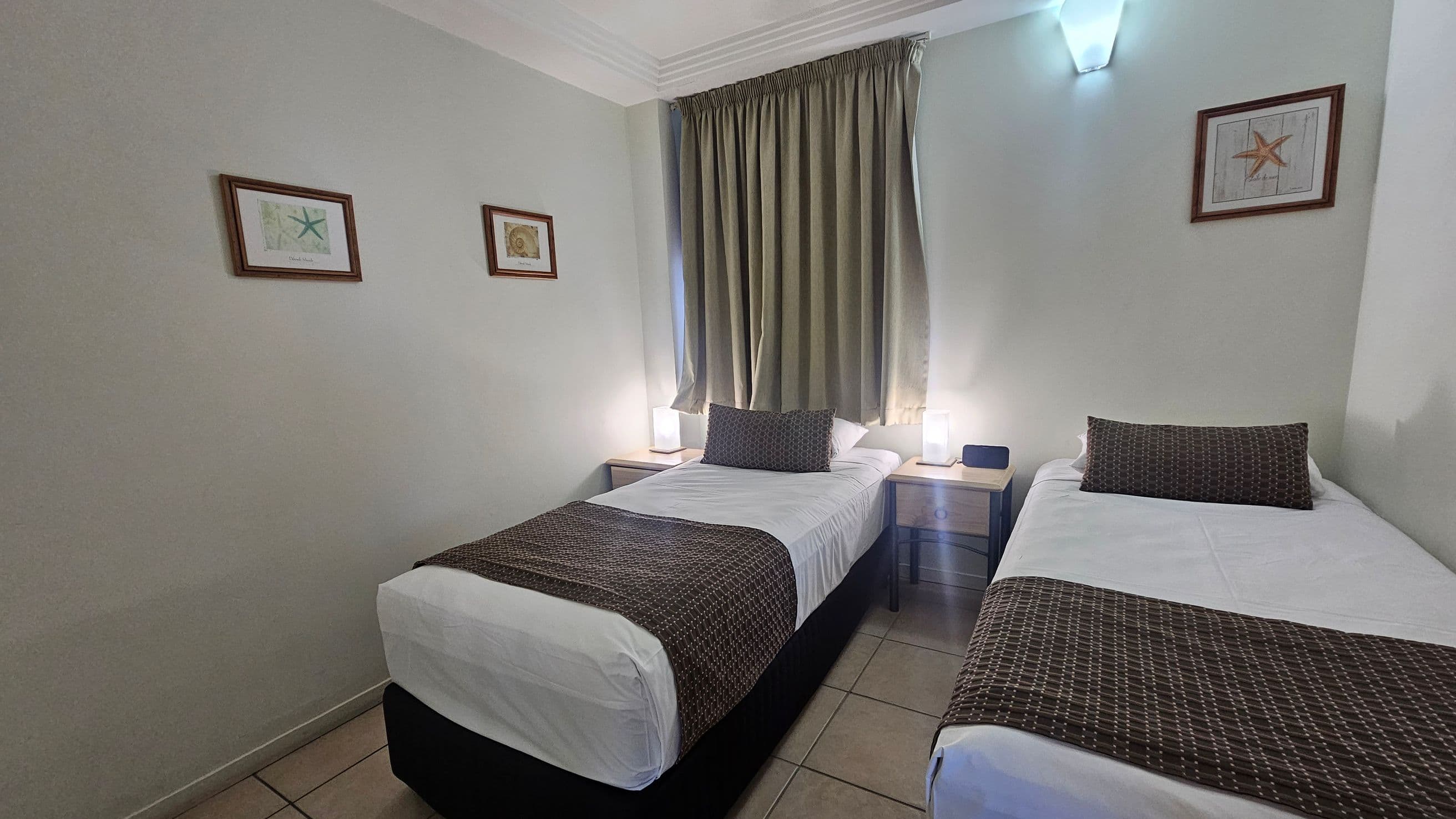 Two single beds with white sheets and dark patterned throws are positioned side by side. Each bed has a matching dark pillow. A small wooden nightstand with a lamp is between the beds. Above the beds, a window with green curtains is centred on the wall. Three framed pictures hang on the walls, depicting starfish and shells. The floor is tiled, and a wall-mounted light fixture is above the window.