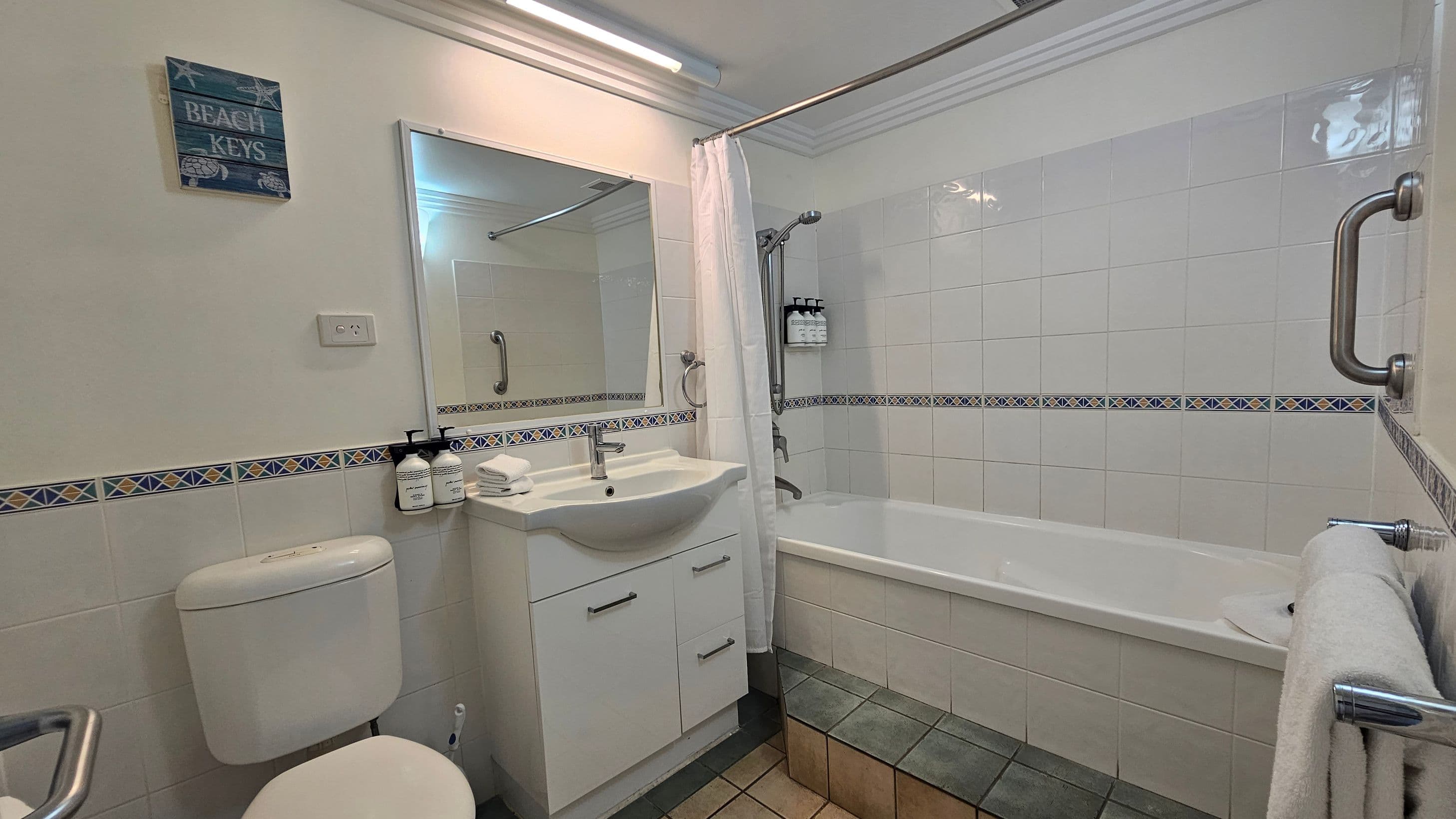A bathroom with a white bathtub on the right, featuring a showerhead and a white curtain. Above the tub are mounted bottles of toiletries. The wall is tiled with a decorative border. On the left, there's a white toilet and a sink with a mirror above it. The sink has a cabinet below and toiletries on the counter. A towel hangs on a rail beside the tub. The floor is tiled in a mix of grey and beige. A small sign on the wall reads "BEACH KEYS."