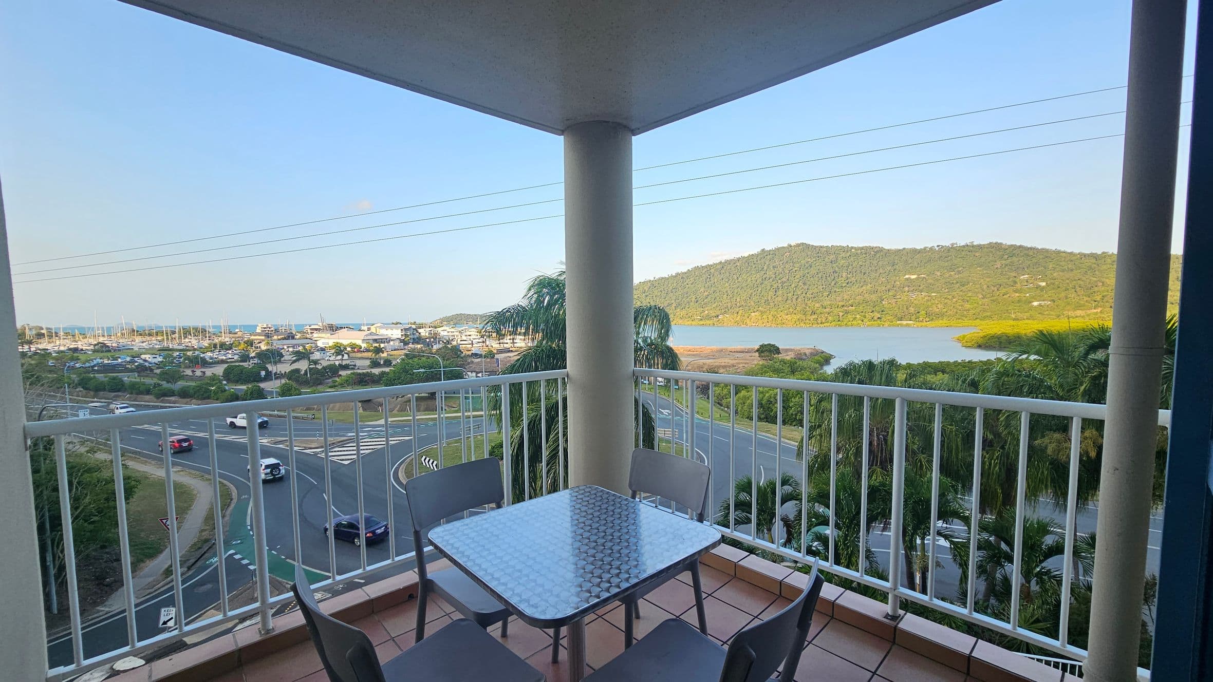 A balcony with a square metal table and four chairs overlooks a scenic view. In the foreground, the balcony railing frames the scene. Below, a road with cars curves around a roundabout. In the background, a marina with boats is visible to the left, and a large hill with greenery rises to the right, bordered by a body of water. Palm trees are scattered throughout the landscape. The ceiling of the balcony is visible, supported by a central pillar.