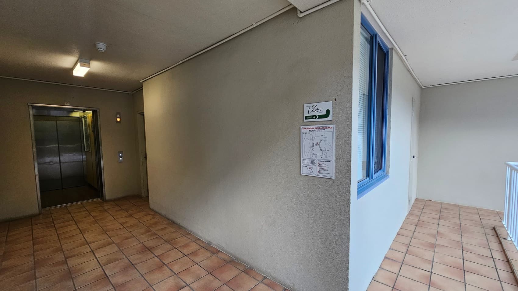 A corridor with a tiled floor and a beige ceiling. On the left, there is an open lift with metallic doors and a control panel beside it. The right side features a wall with a blue-framed window and a door. A sign with "Vista" and "Unit 3" is mounted on the wall, along with an evacuation diagram. The corridor extends to a railing on the far right.