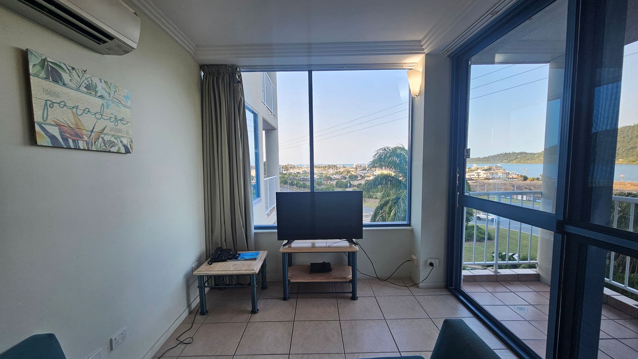 A room with a tiled floor and a large window offering a view of a marina and distant hills. In the centre, a television sits on a small table. To the left, a wall-mounted air conditioner is above a decorative sign with tropical designs. A small side table holds a few items. On the right, a glass door leads to a balcony. The ceiling has crown moulding, and a curtain partially covers the window.