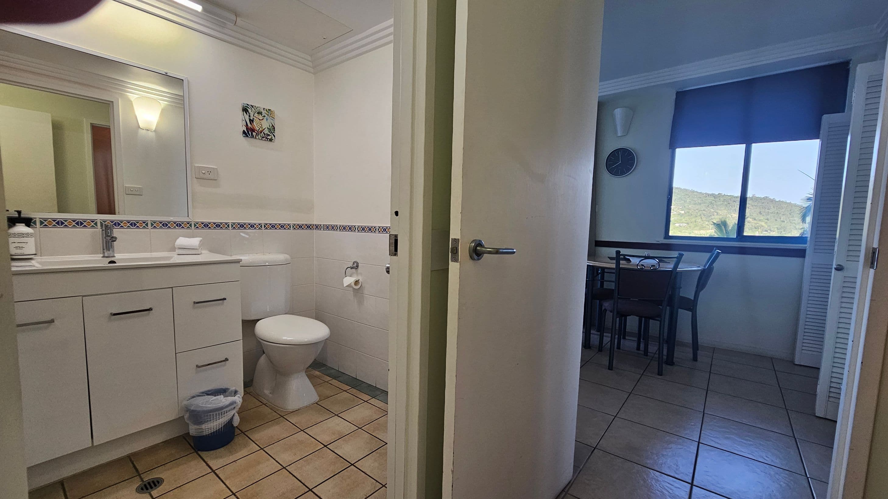 A bathroom on the left with a large mirror above a sink, a toilet, and a small waste bin on a tiled floor. The wall has a decorative tile border. On the right, a dining area with a table and chairs is visible through a partially open door. A large window shows a view of greenery outside. A clock is mounted on the wall above the table. The floor is tiled, and the ceiling has crown moulding.