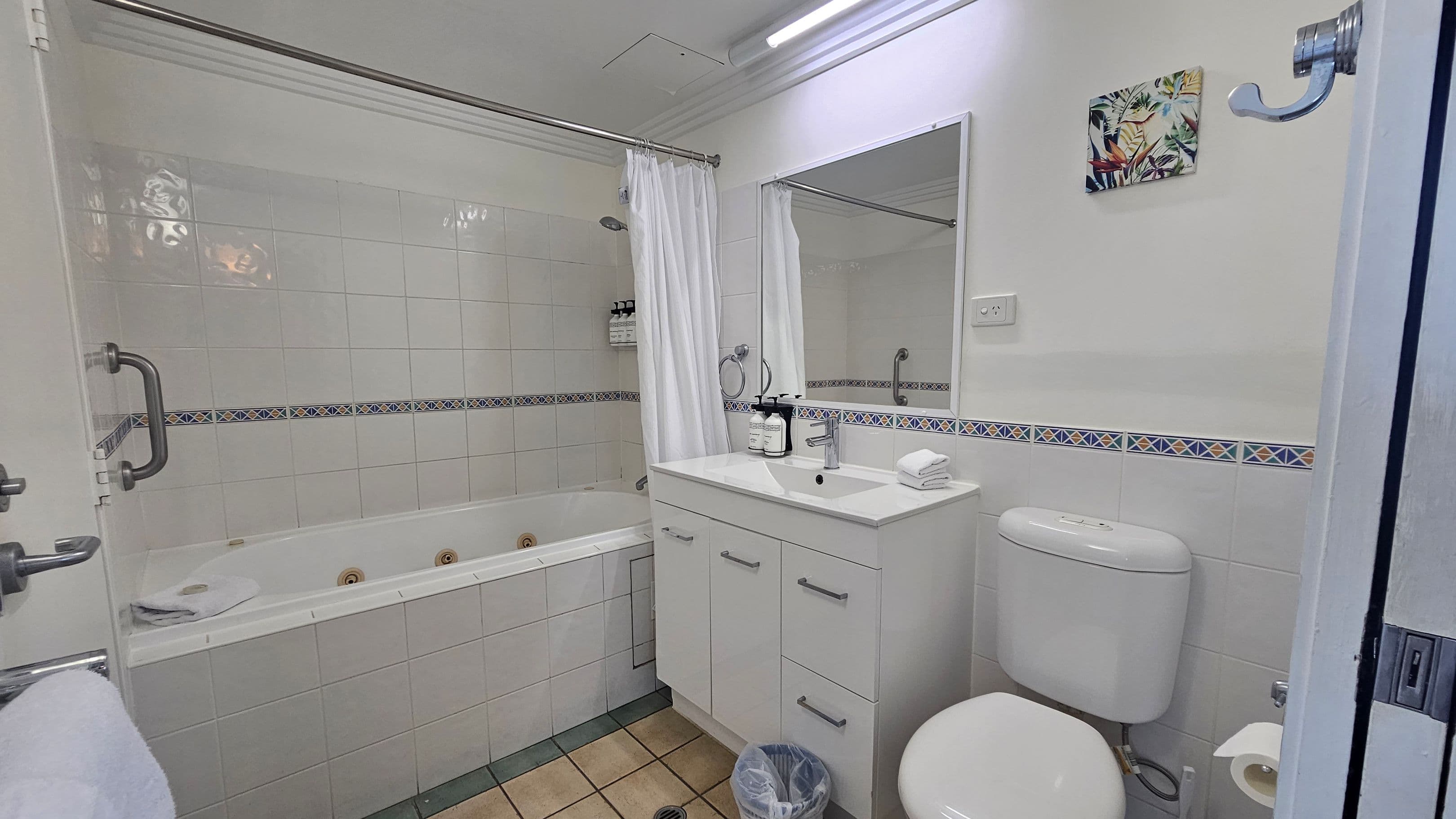 A bathroom with a white tiled floor and walls. On the left, there's a bathtub with a showerhead and a white curtain. The bathtub has a grab bar and toiletries on a shelf. In the centre, a white vanity with a mirror above it holds soap dispensers and a folded towel. On the right, there's a toilet with a roll of toilet paper nearby. Above the toilet, a small colourful painting is mounted on the wall. A metal hook is visible on the right side.