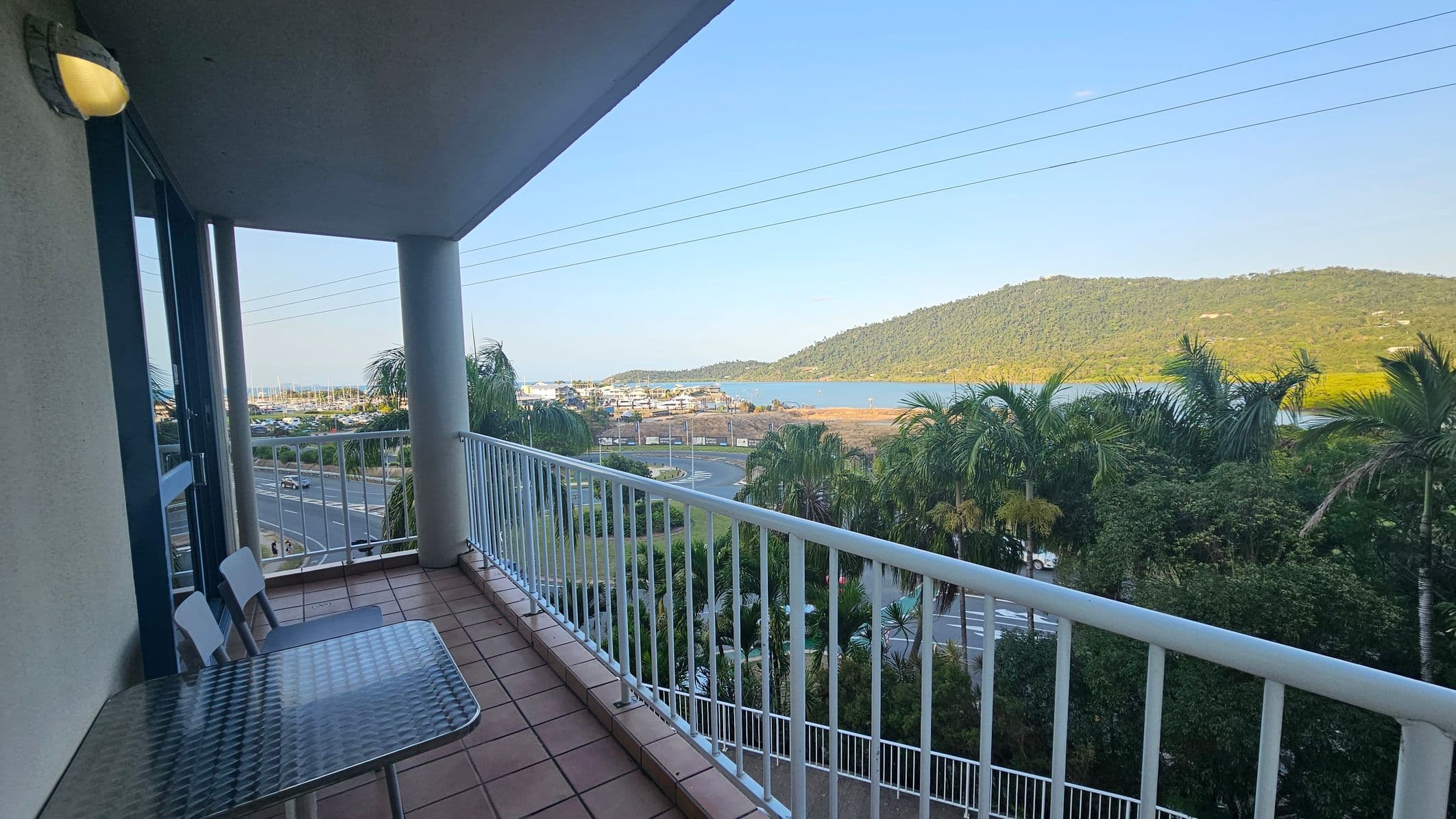 A balcony with a tiled floor and a metal table with two chairs on the left. The railing runs along the edge, overlooking a road and lush greenery with palm trees. In the distance, a body of water is visible, bordered by a green hill. A light fixture is mounted on the wall to the left.