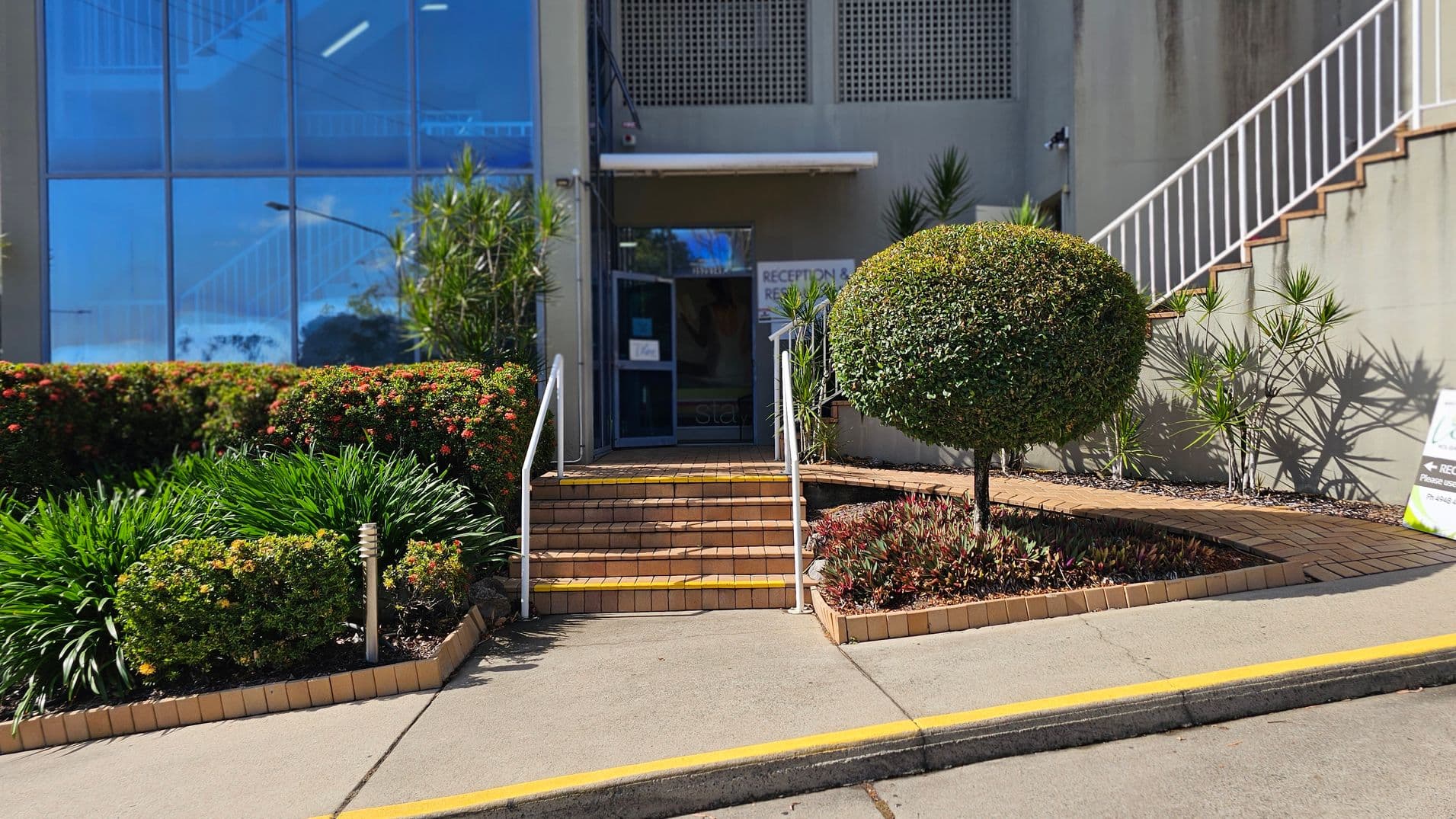 A building entrance with a set of brick steps leading up to a glass door. On the left, there are neatly trimmed bushes and flowering plants in a raised garden bed. On the right, a round, manicured shrub is surrounded by smaller plants. A white railing lines the steps, and a sign is visible near the entrance. The building has a large glass window reflecting the surroundings, and a staircase with a white railing is on the right side. The pavement has a yellow line along the edge.