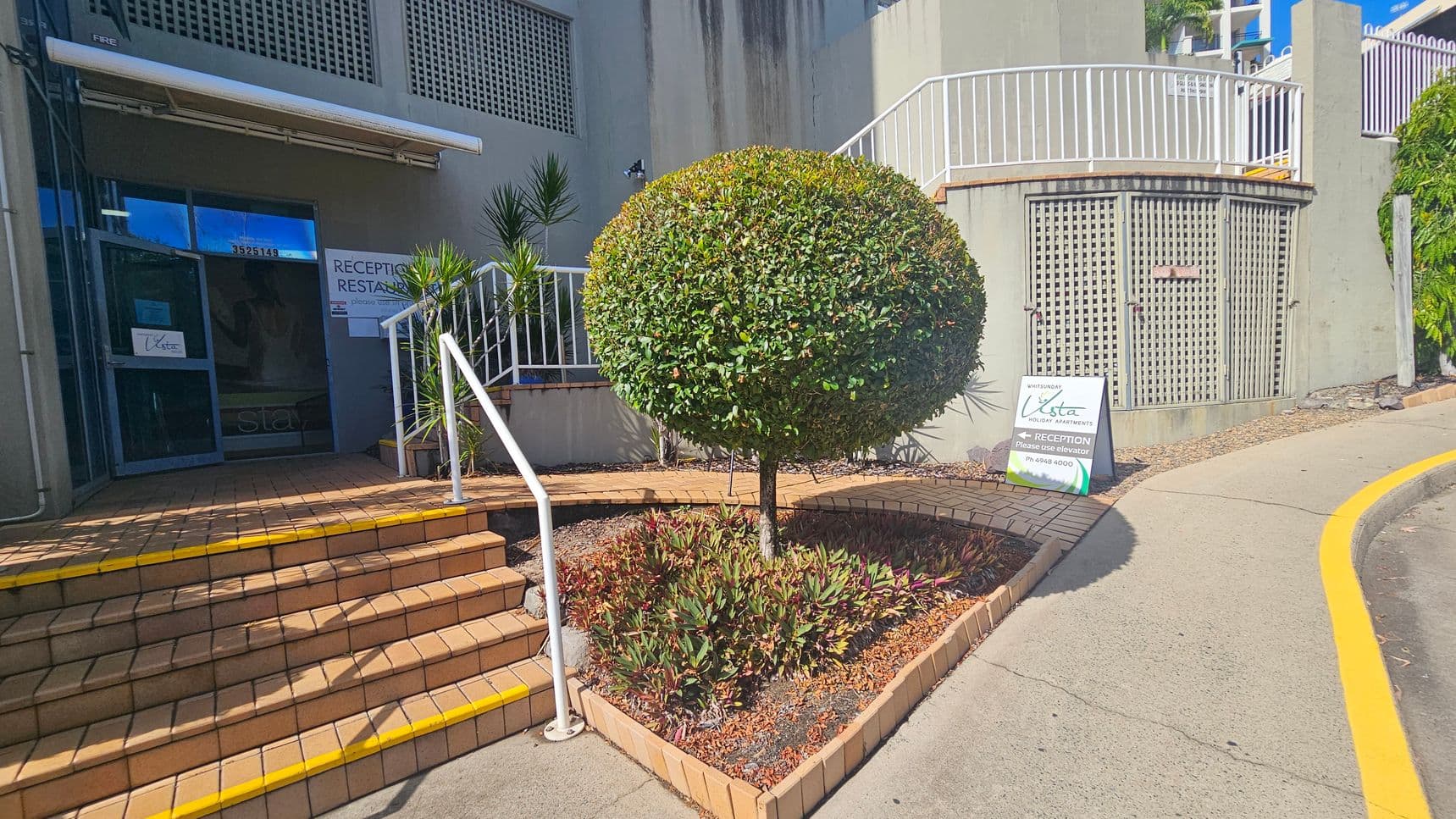 A building entrance with a blue-framed glass door on the left, marked with signs for reception and restaurant. Brick steps with yellow edges lead up to the door, flanked by white railings. In the centre, a neatly trimmed round bush is surrounded by a small garden bed with various plants. On the right, a curved concrete path with a yellow line leads past a sign for "Vista Holiday Apartments" and a green lattice structure.