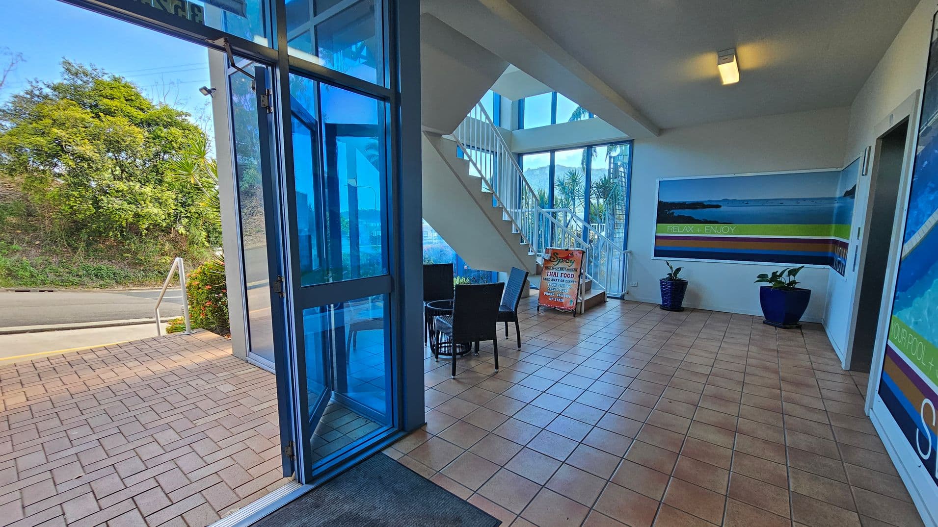 Inside a building with a tiled floor and a white ceiling. On the right, there are two large posters with scenic images and the words "RELAX + ENJOY." Two blue plant pots are placed beneath the posters. In the centre, a staircase with white railings leads upwards. A small seating area with dark chairs and a table is nearby. On the left, a glass door opens to the outside, showing a paved area and greenery. A sign advertises Thai food near the stairs.