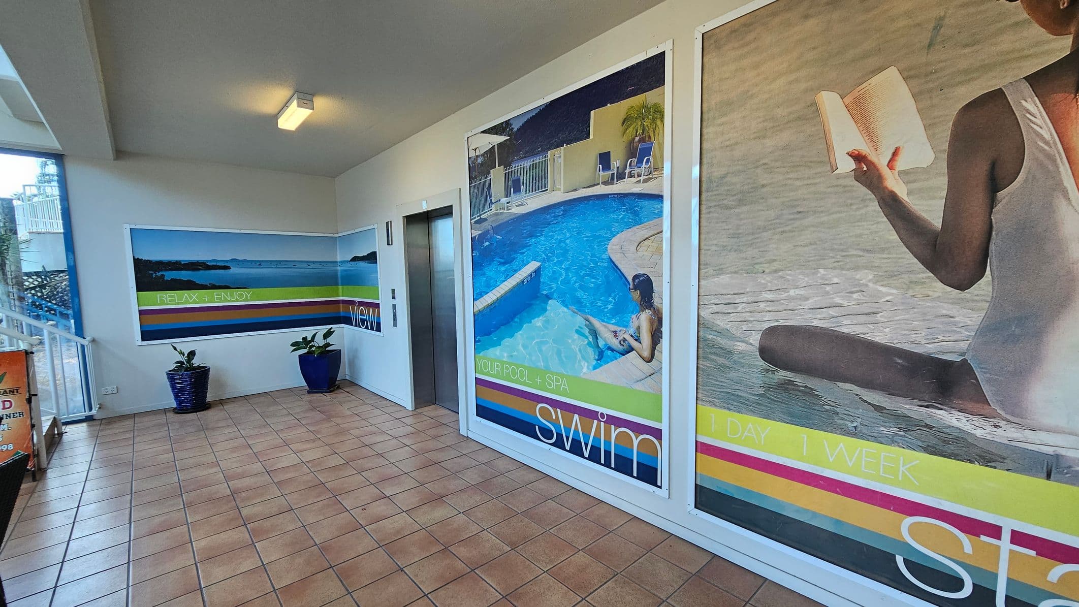 A hallway with tiled flooring and a white ceiling featuring a fluorescent light. On the right, there are large posters; one shows a woman sitting by a pool, and another depicts a person reading by the water. In the centre, there's an elevator with metallic doors. On the left, a poster displays a scenic view with the words "RELAX + ENJOY." Two blue plant pots with green plants are placed against the wall. A glass window and railing are visible on the far left.