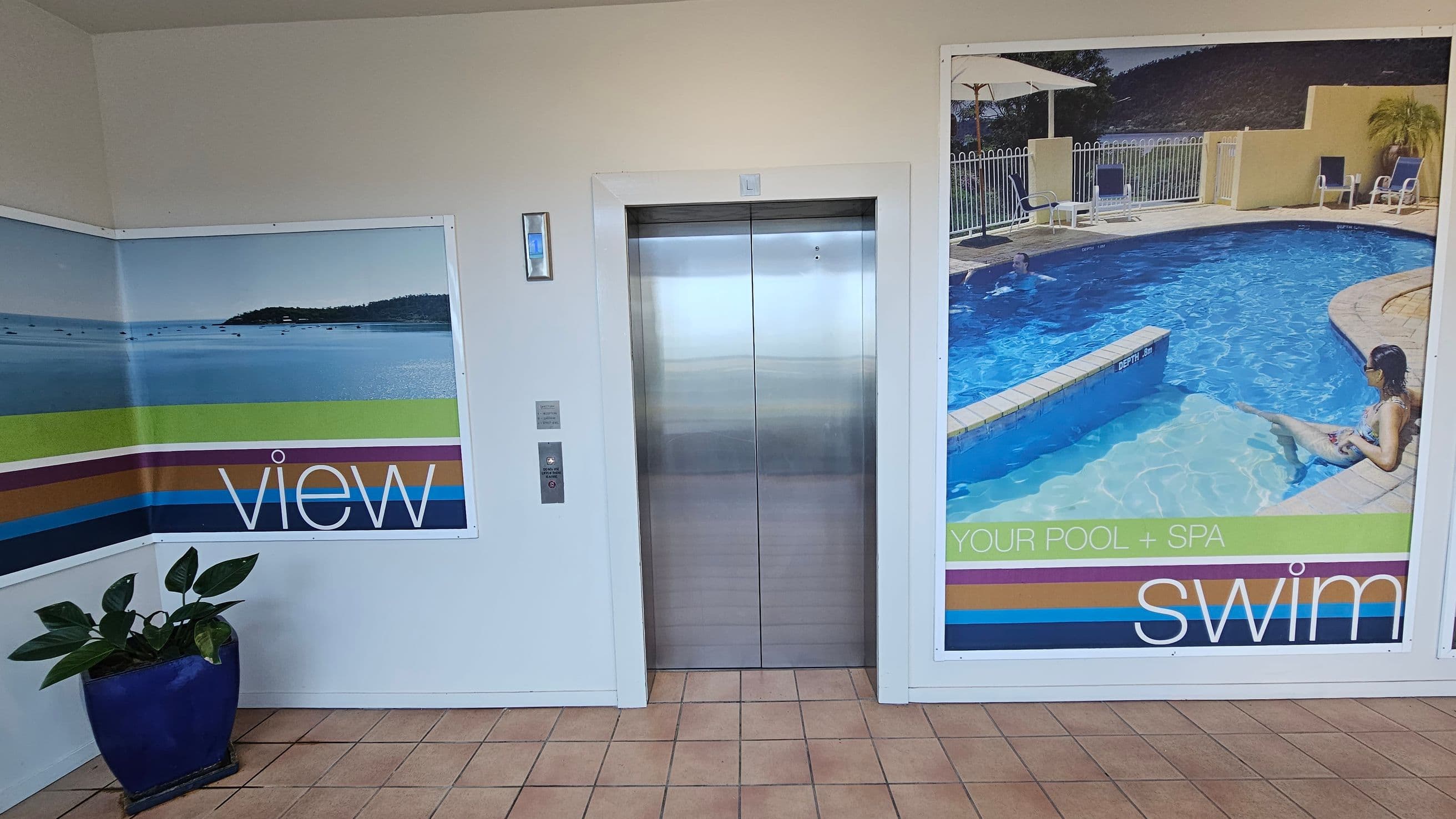 A tiled floor leads to a central lift with metallic doors. On the left, a large poster shows a serene ocean view with the word "view" in bold white letters. Below it, a blue pot holds a leafy green plant. On the right, another poster displays a swimming pool scene with a person relaxing by the water, accompanied by the words "YOUR POOL + SPA" and "swim" in white letters. The walls are light-coloured, and the ceiling is plain.