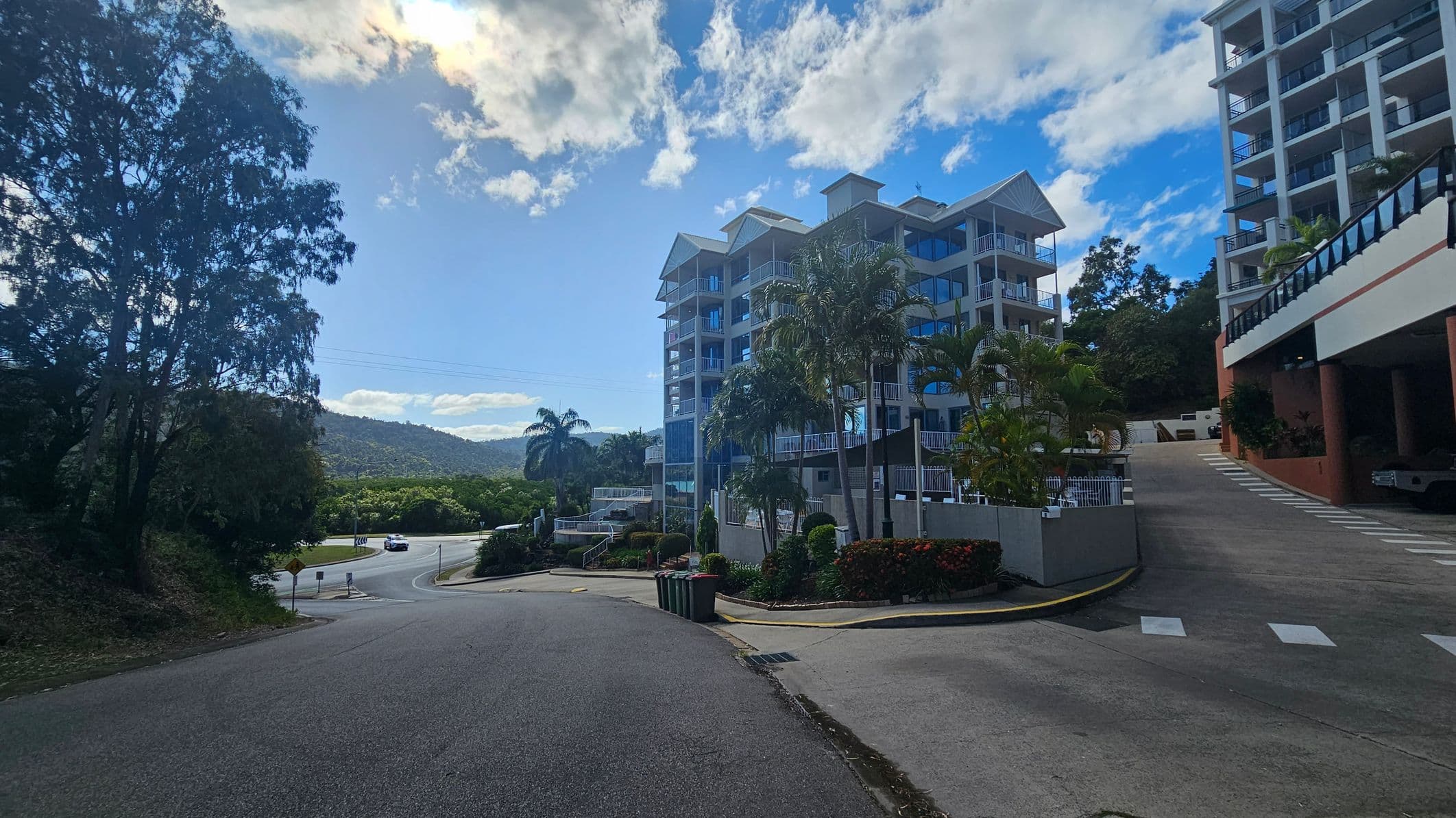 On the left, a large tree stands beside a road that curves into the distance, with a car approaching a roundabout. In the centre, a multi-storey building with balconies is surrounded by palm trees and shrubs. On the right, another building with a sloped driveway leading upwards. The foreground features a paved road with painted lines and a few bins near the kerb. In the background, hills are visible under a partly cloudy sky.