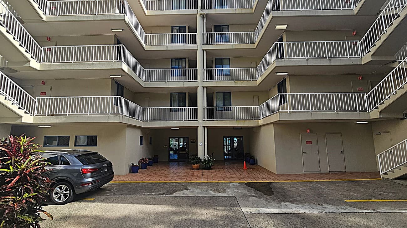 A multi-storey building with balconies featuring white railings. The ground level has a tiled entrance with potted plants and glass doors. A grey car is parked on the left, next to some foliage. The building's exterior is light-coloured, and there are visible staircases on the right. The ground is a mix of concrete and painted surfaces.